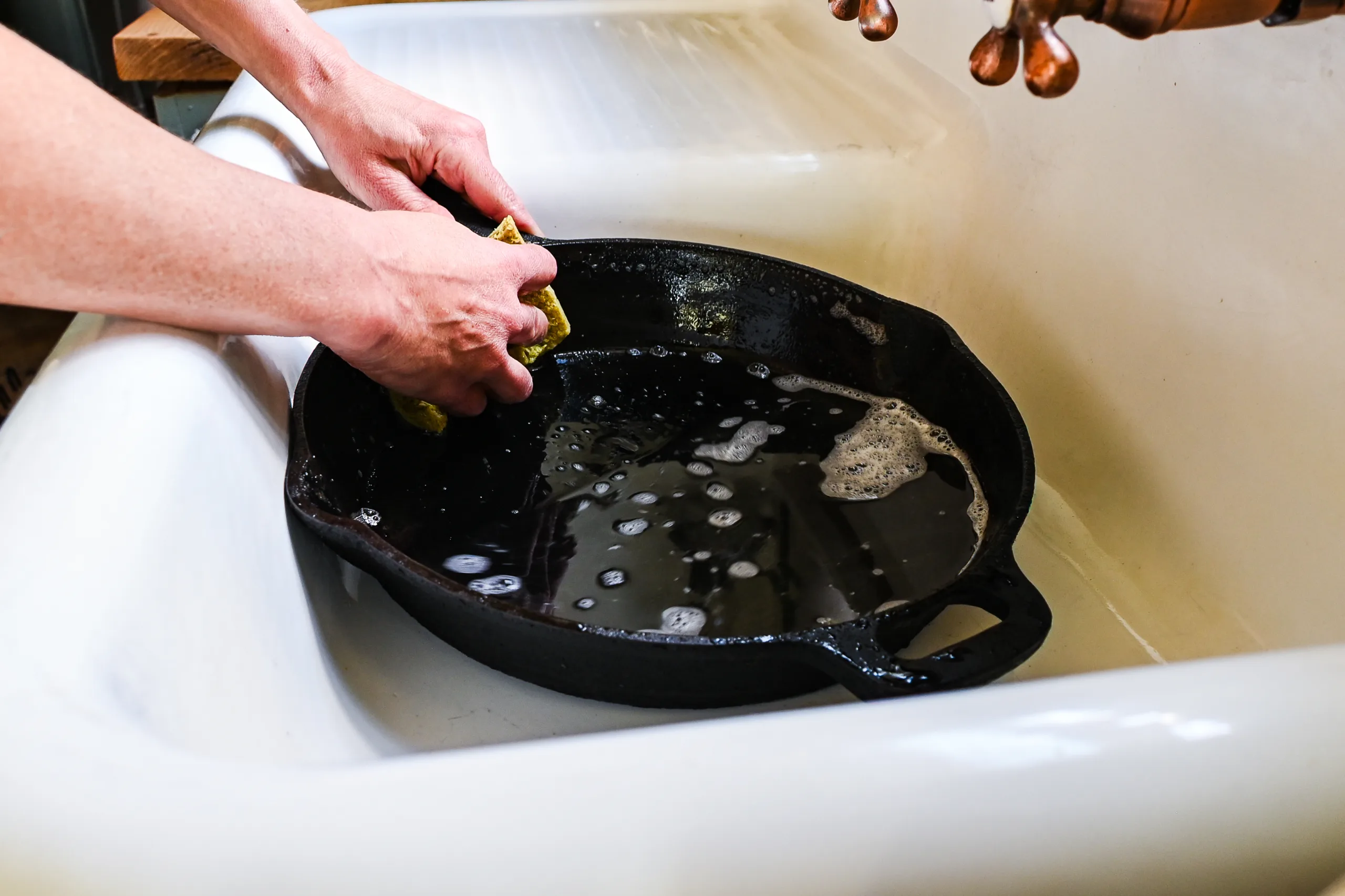 Hands washing a cast iron skillet with a sponge in a sink