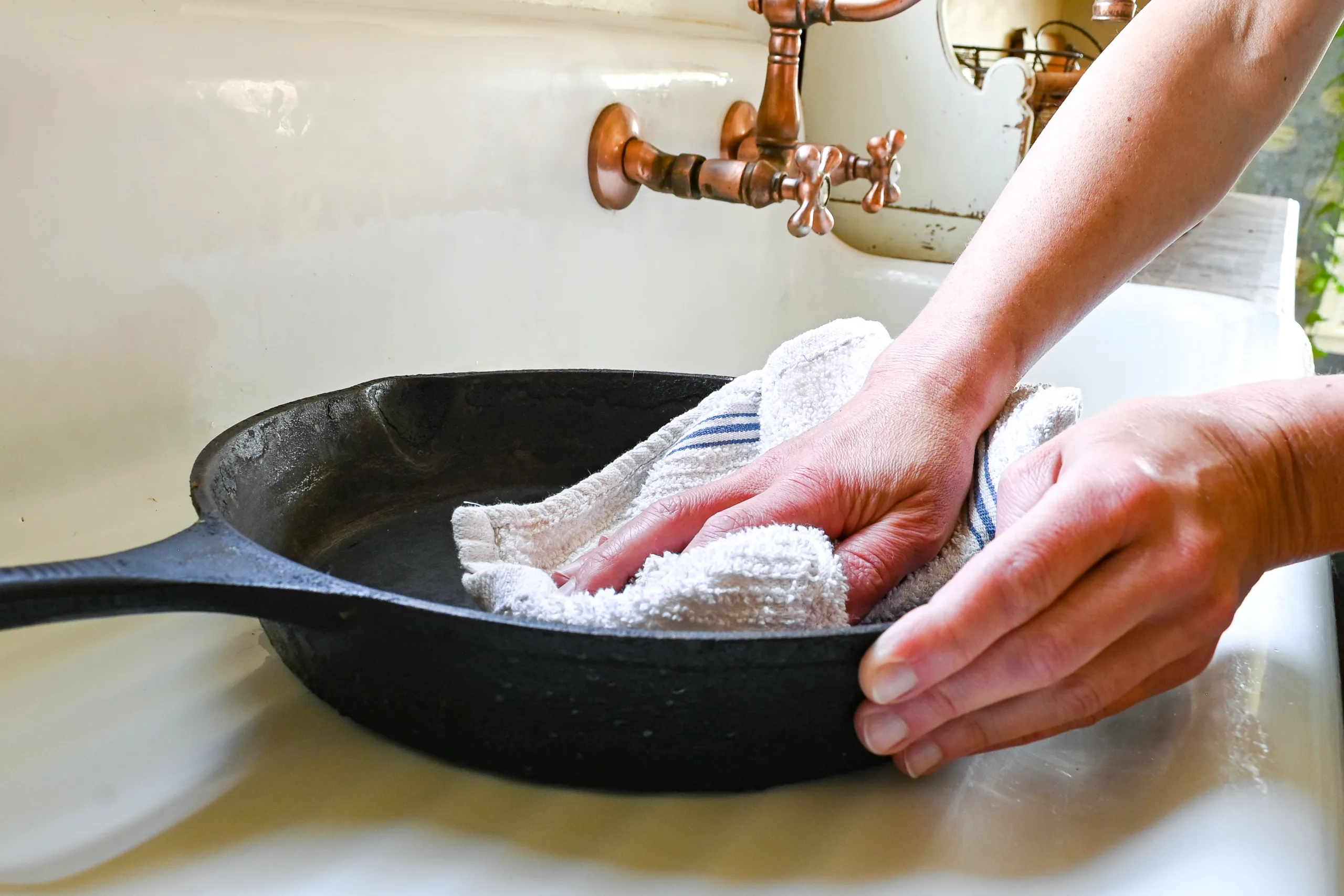 Hands drying a cast iron skillet with a towel in a farmhouse sink
