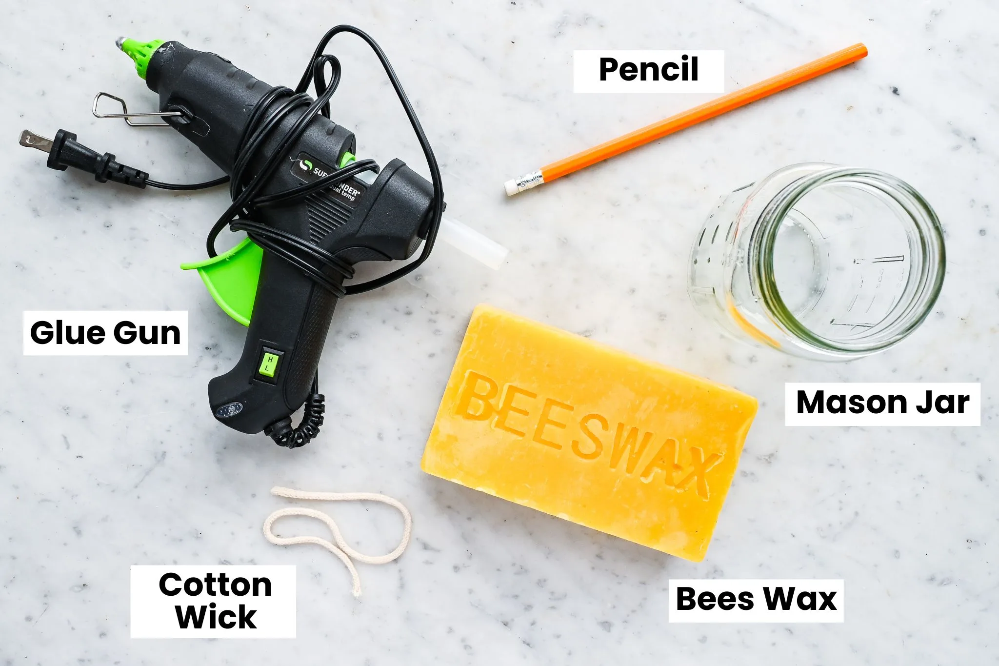 Supplies for making beeswax candles in a mason jar, including beeswax, wick, hot glue gun, pencil, and glass jar
