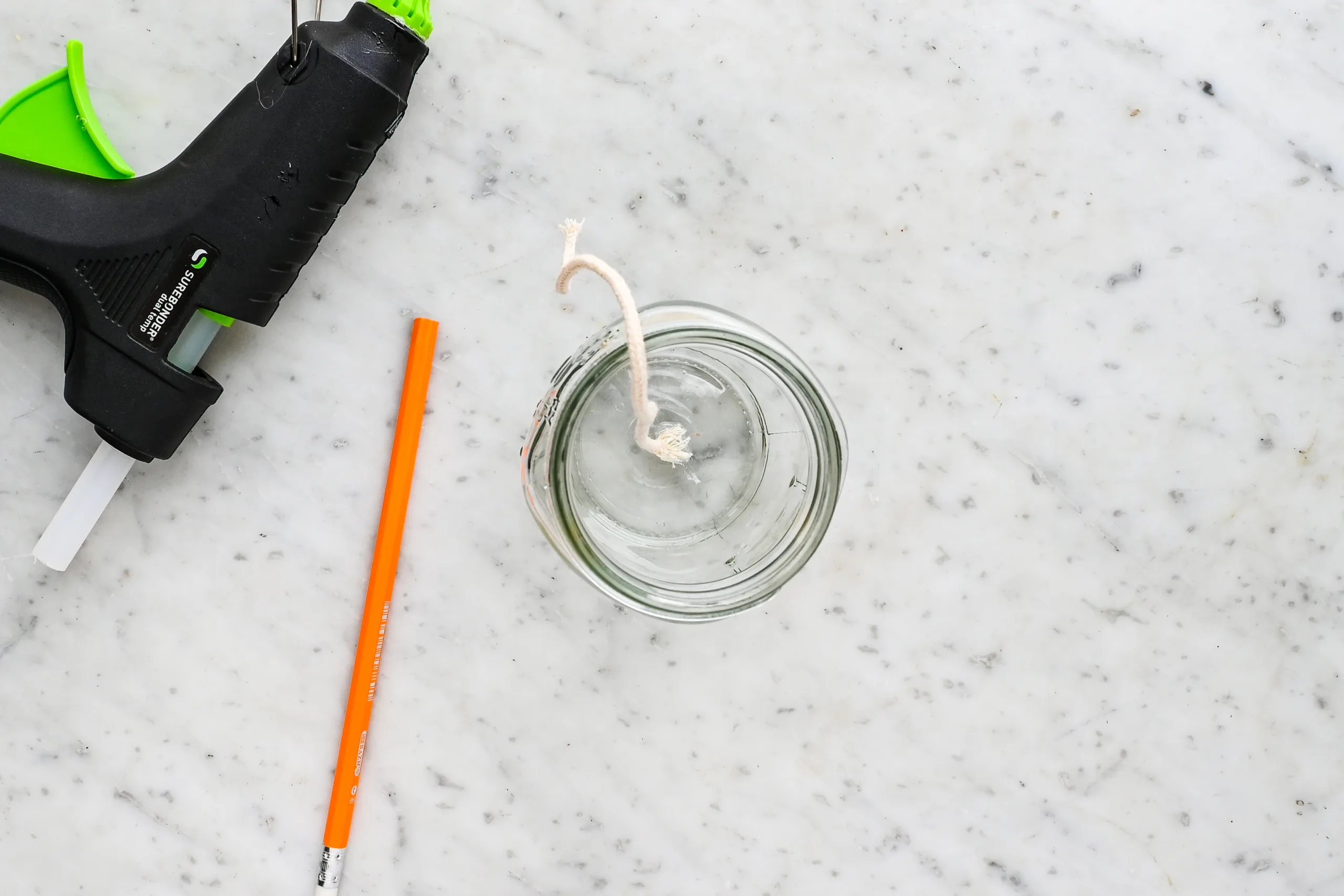 Candle wick glued to the bottom center of a mason jar with a hot glue gun and pencil nearby