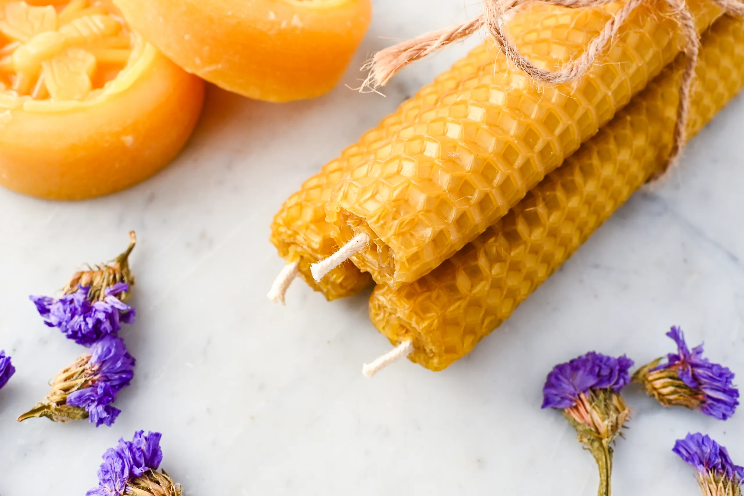 Three finished rolled beeswax candles tied with twine, with dried purple flowers and beeswax in the background