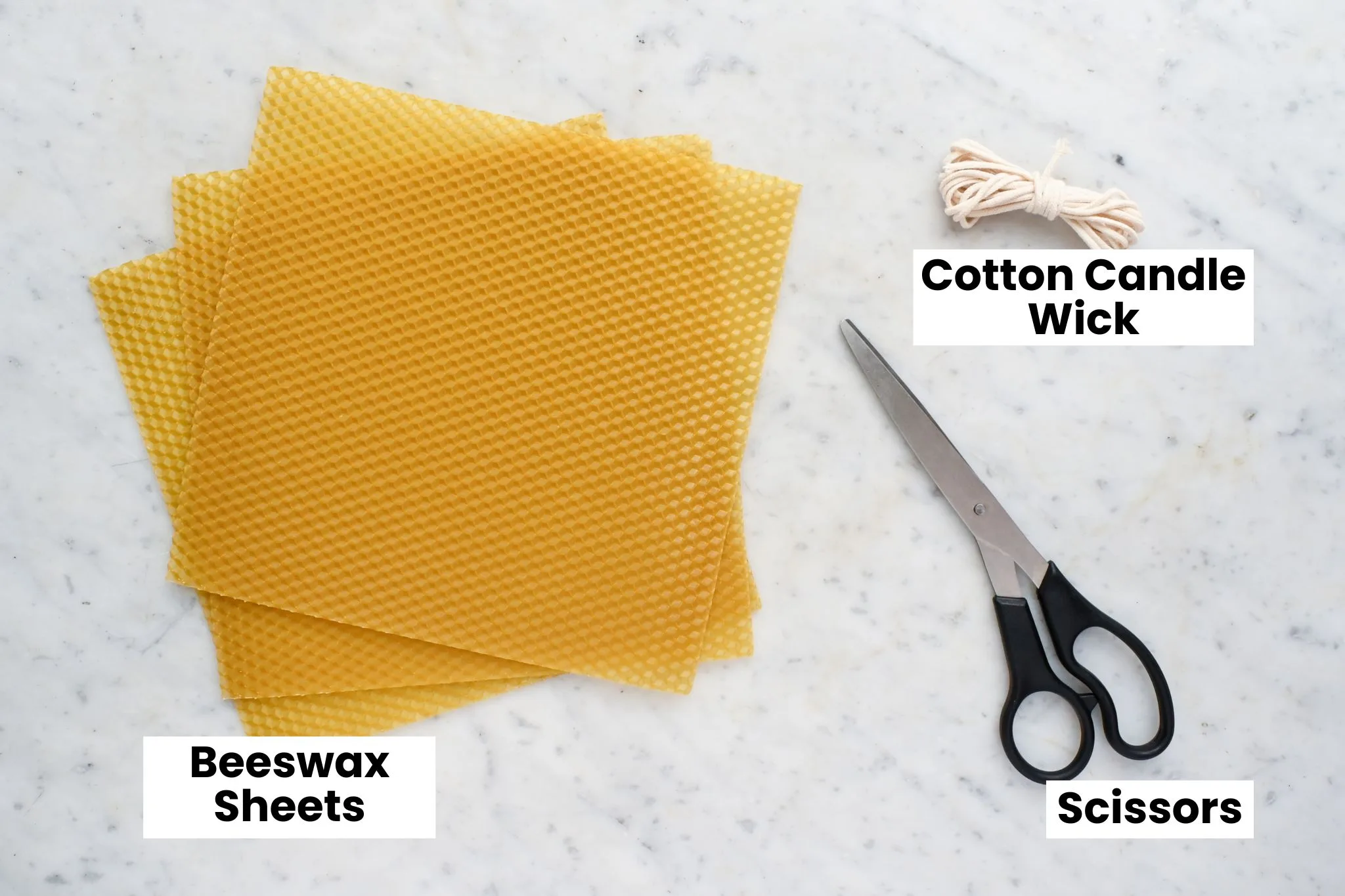 Supplies for making old-fashioned rolled beeswax candles labeled on a marble surface, including beeswax sheets, cotton candle wick, and scissors