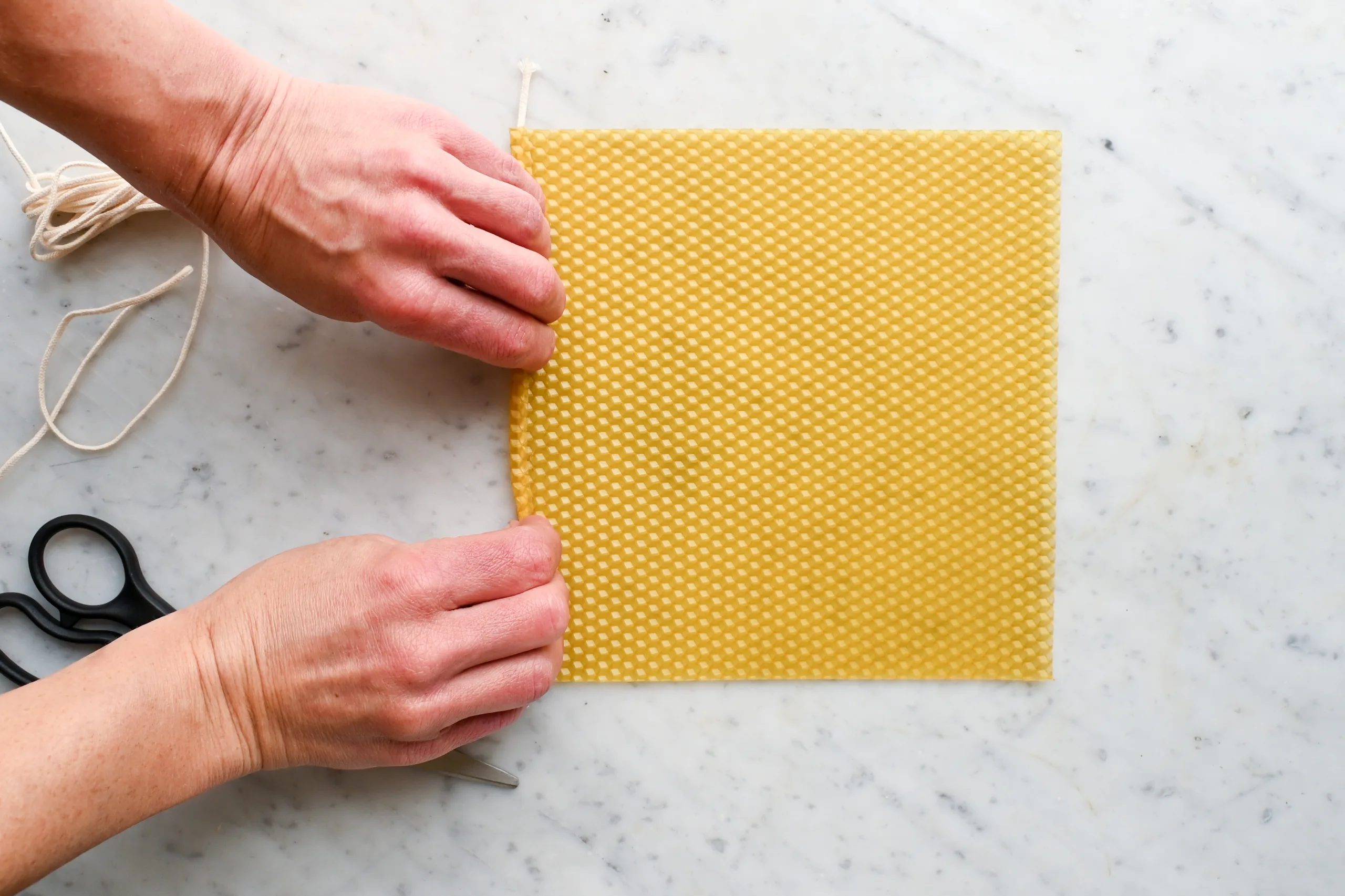Hands pressing the edge of a beeswax sheet around a wick to begin rolling a beeswax candle