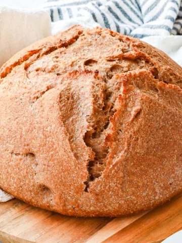 Fresh milled spelt sourdough bread with a rustic golden crust cooling on a wooden board beside a striped kitchen towel.