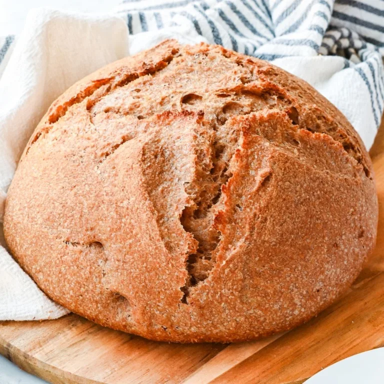 Fresh milled spelt sourdough bread with a rustic golden crust cooling on a wooden board beside a striped kitchen towel.