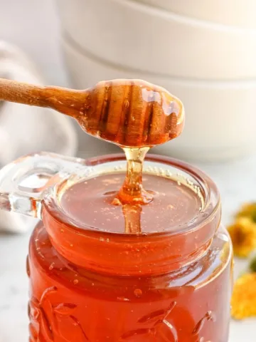 Homemade dandelion honey dripping from a wooden honey dipper into a glass jar.