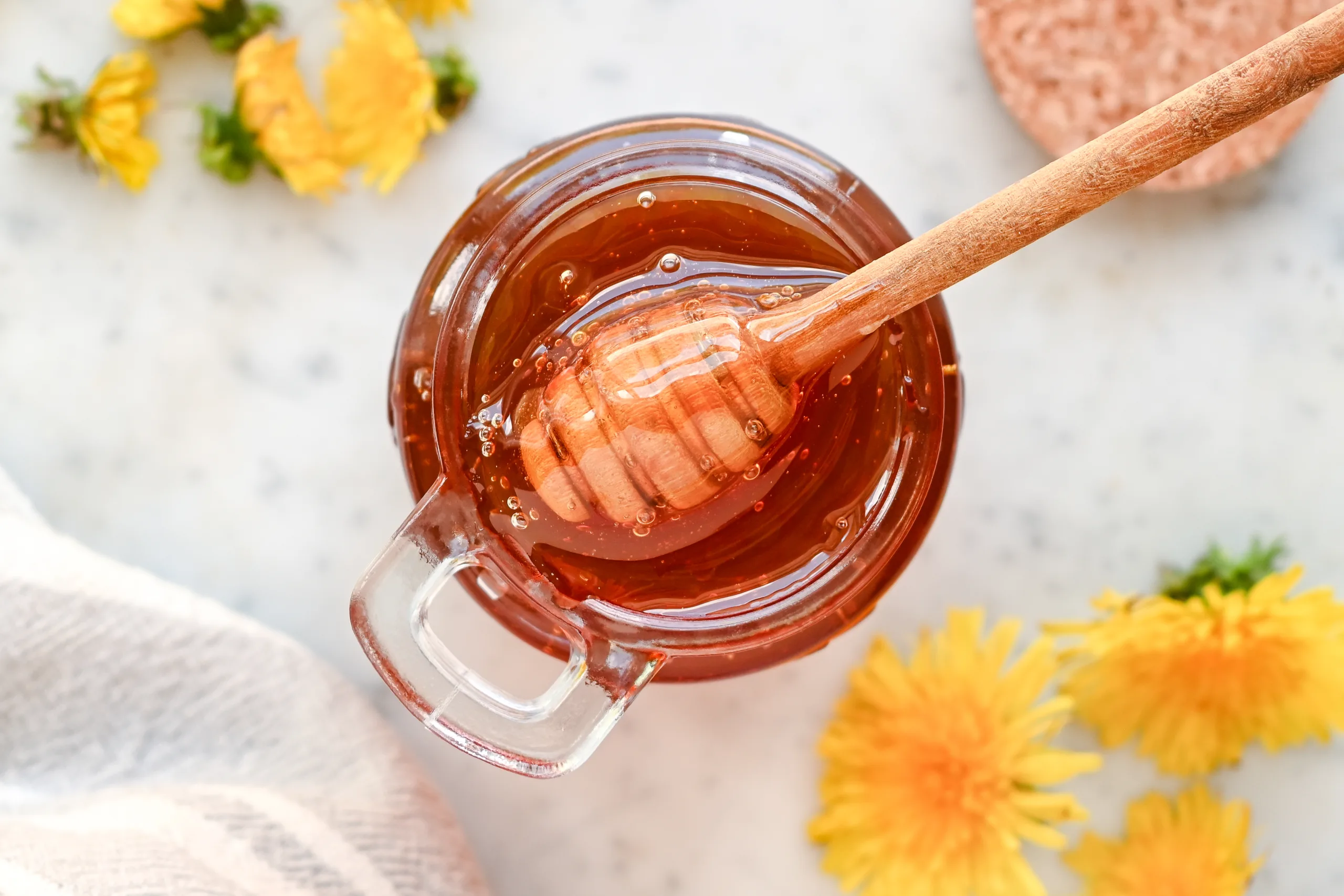 Golden dandelion honey in a glass jar with a honey dipper and dandelion flowers.