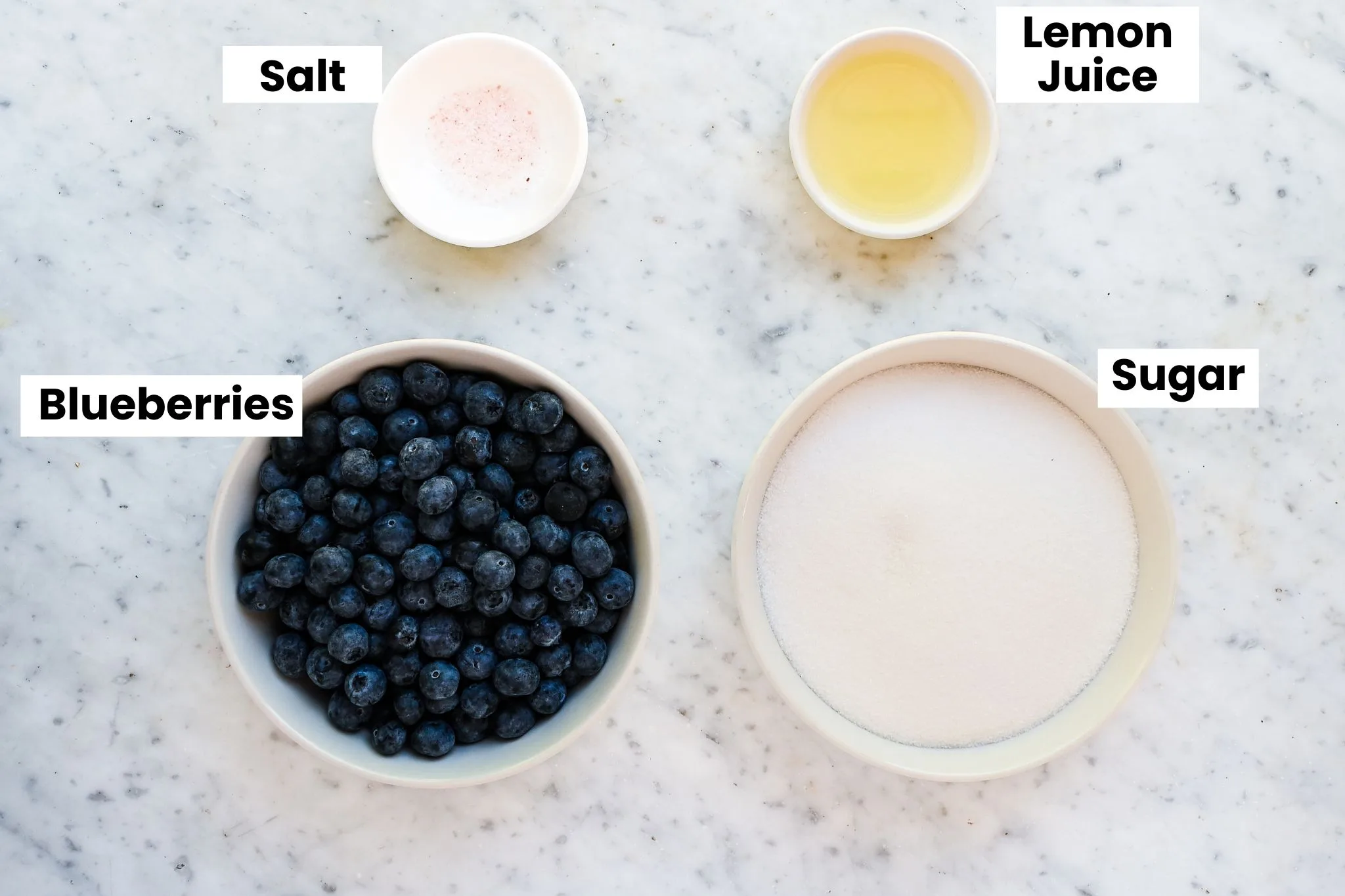 Overhead view of ingredients for homemade blueberry jam without pectin, including fresh blueberries, granulated sugar, lemon juice, and salt arranged in white bowls on a marble countertop.