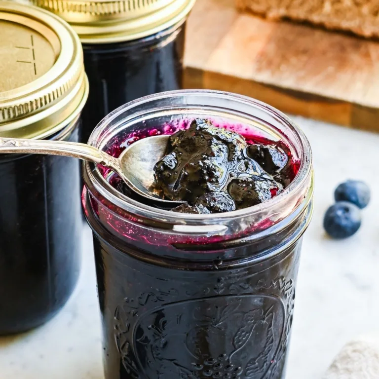 Rustic mason jar filled with thick homemade blueberry preserves beside a vintage spoon and fresh homemade bread, creating a simple old fashioned kitchen scene.