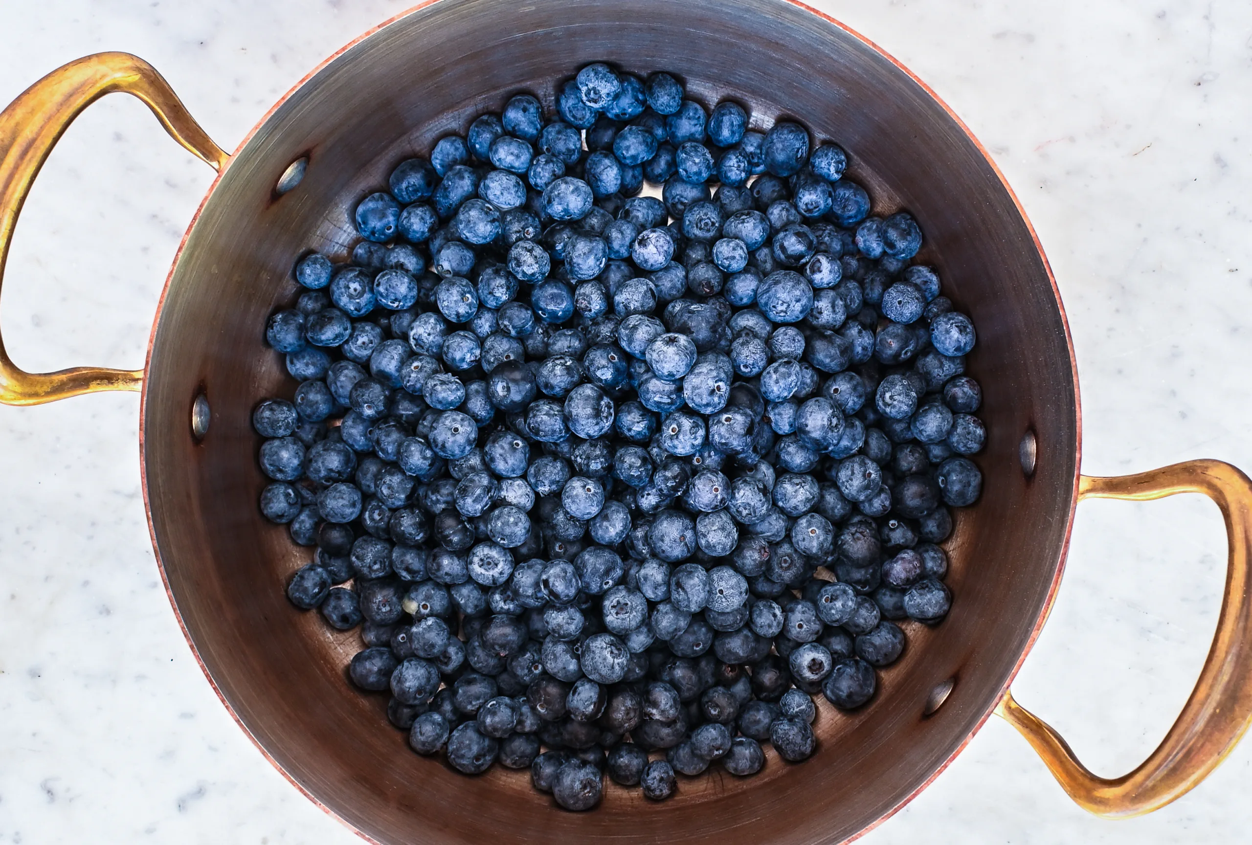 Fresh blueberries filling a copper pot with brass handles on a marble countertop, ready to be cooked into homemade blueberry jam without pectin.