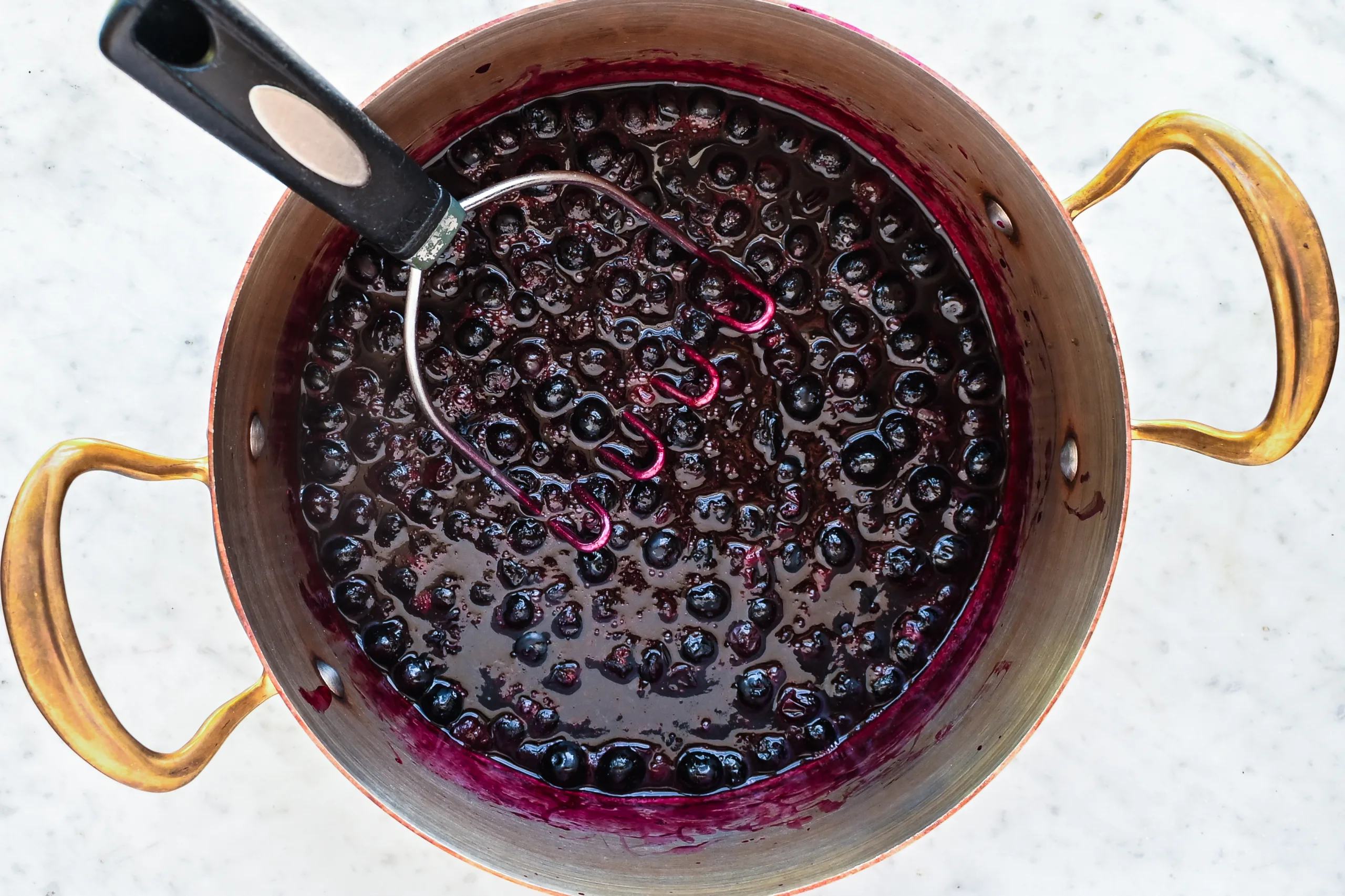 Cooked blueberries simmering in a copper pot while being mashed with a potato masher during the process of making homemade blueberry jam without pectin.