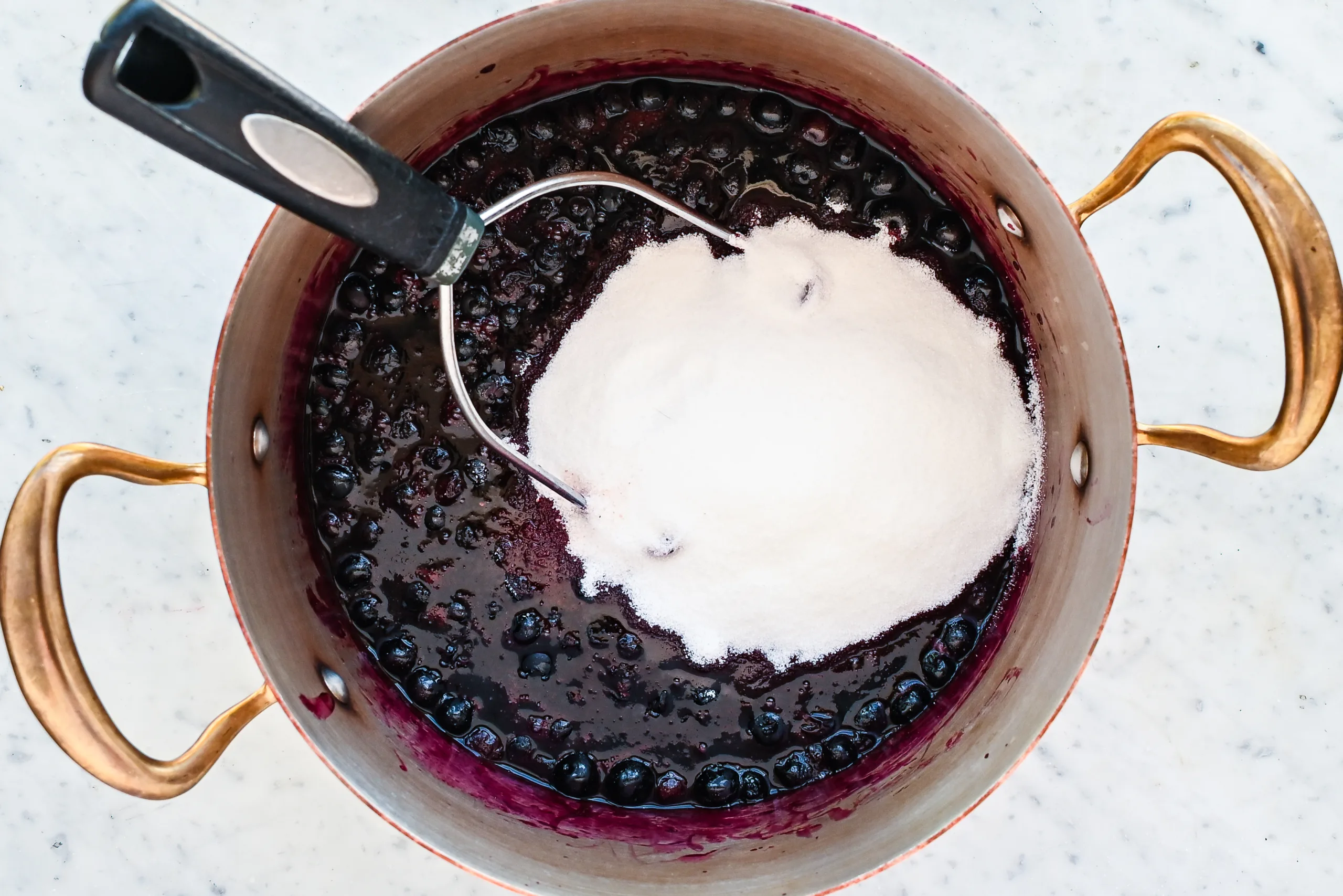 Granulated sugar poured over simmering blueberries in a copper pot with a potato masher resting inside, showing the early cooking stage of homemade blueberry preserves.
