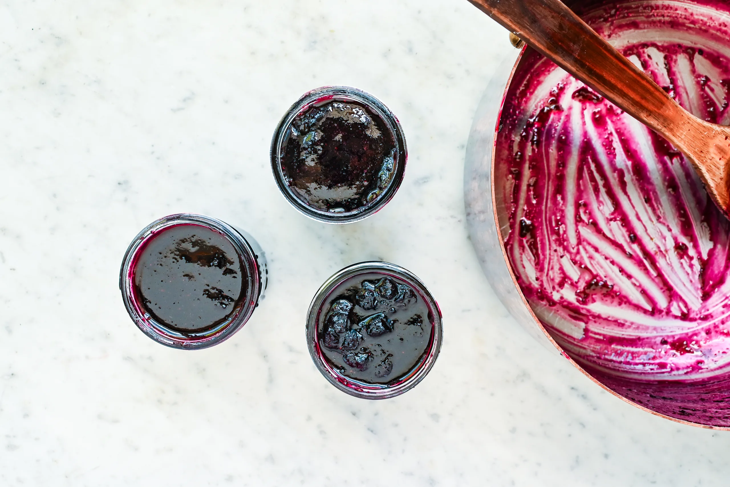 Overhead view of three small jars filled with homemade blueberry jam beside a saucepan streaked with deep purple blueberry mixture and a wooden spoon on a marble countertop.