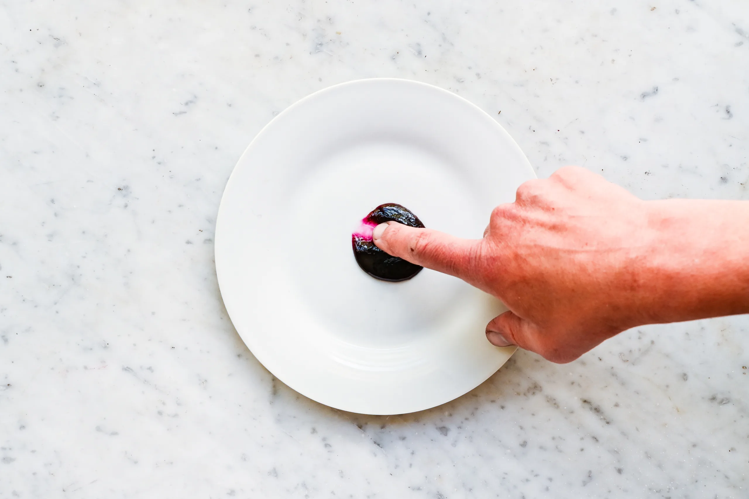 Finger running through a spoonful of homemade blueberry jam on a white plate to test the thickness and set of the jam during cooking.