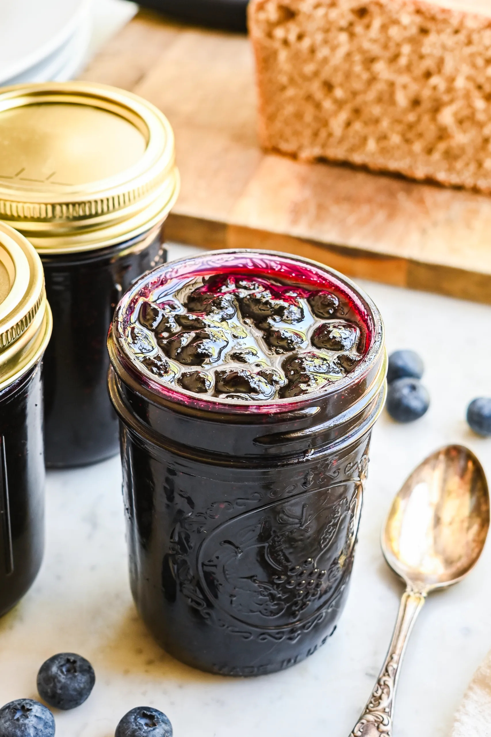 Homemade blueberry jam without pectin in a glass mason jar, surrounded by fresh blueberries, an antique spoon, and slices of homemade bread on a rustic marble countertop.