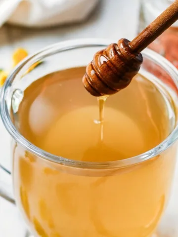 Glass mug of roasted dandelion root tea with honey being drizzled in from a wooden honey dipper.