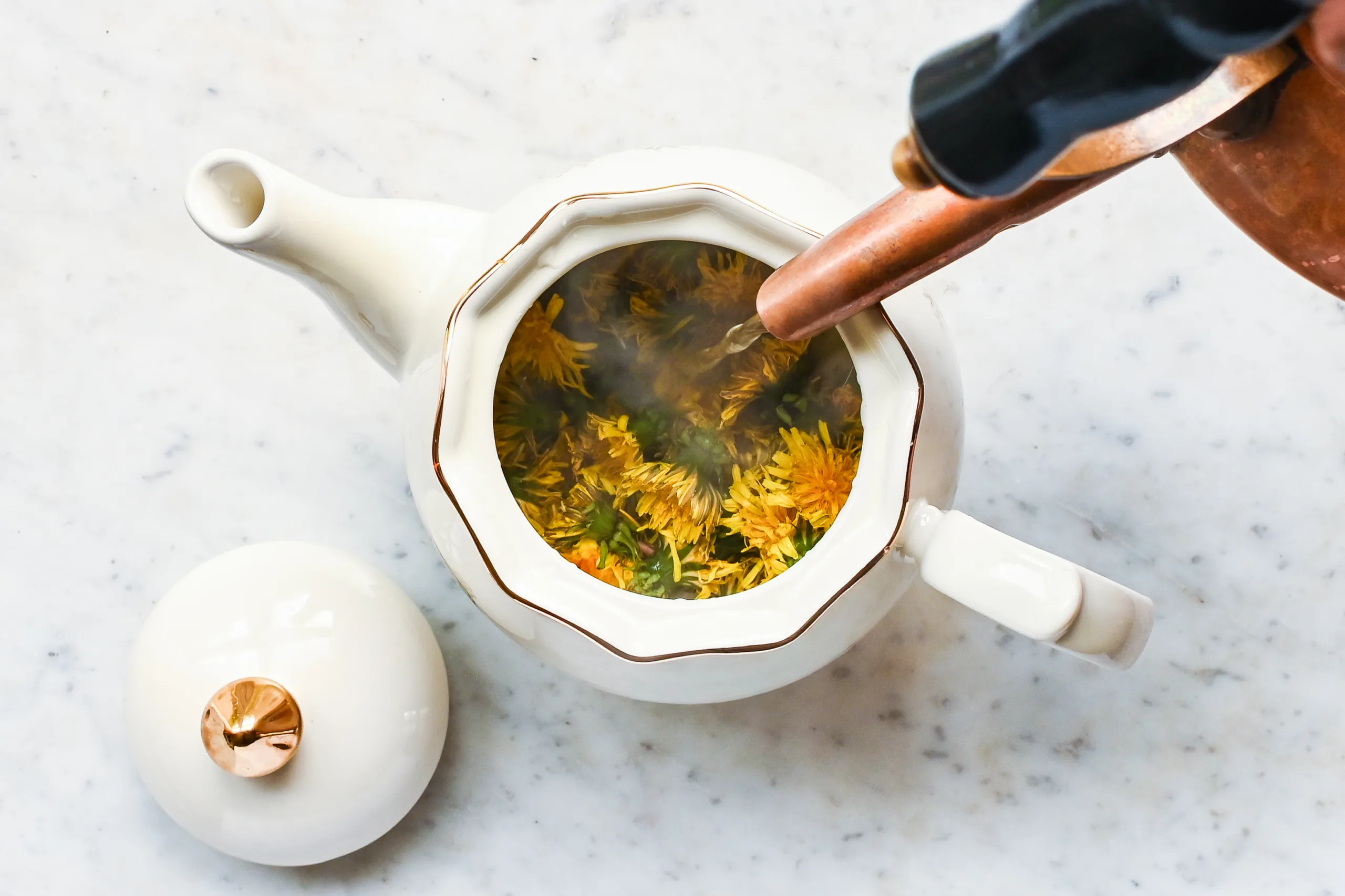 Hot water being poured over fresh dandelion flowers in a white teapot.