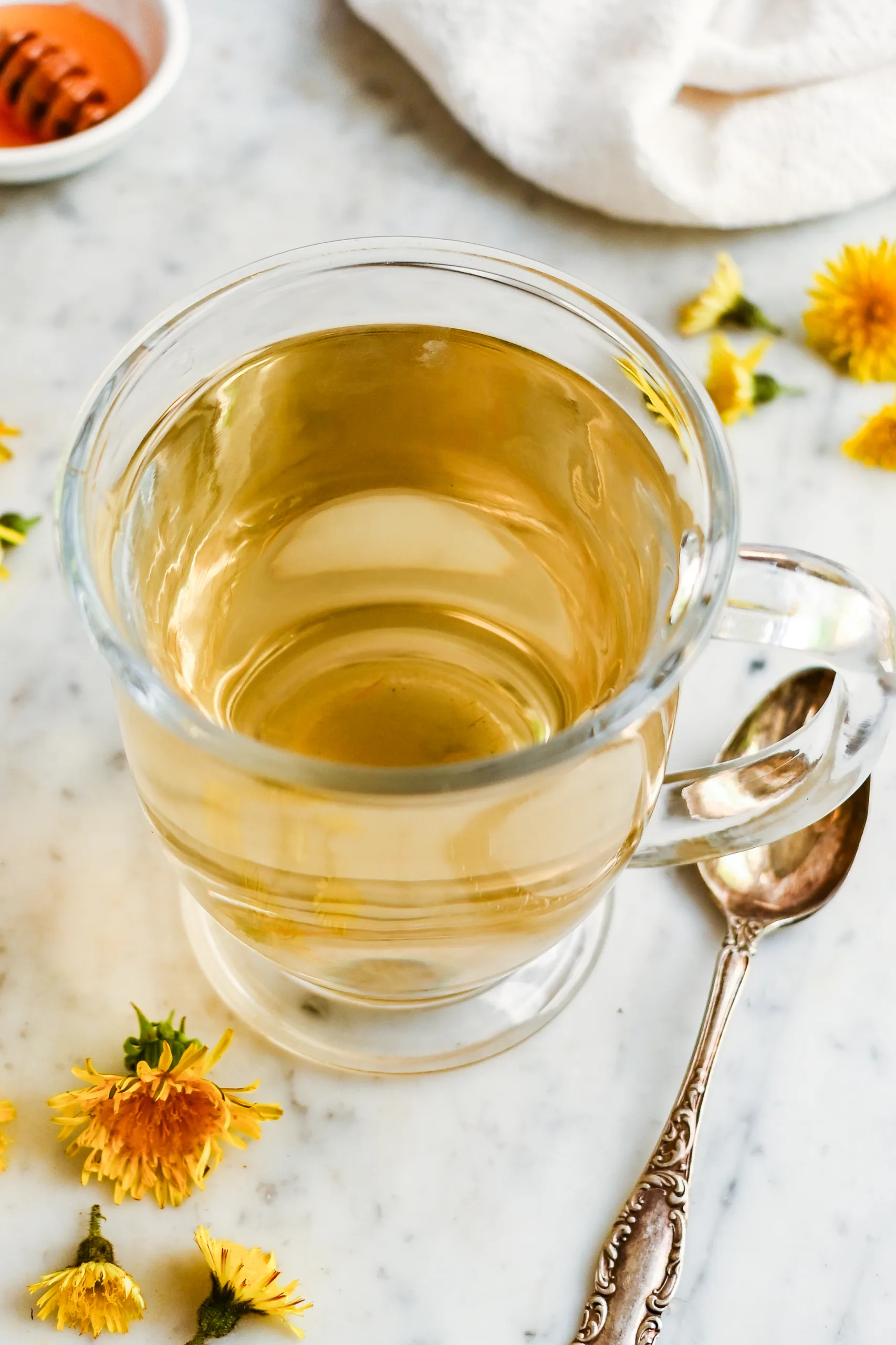 Glass mug of homemade roasted dandelion root tea on a marble counter with dandelion flowers, honey, and a spoon nearby.