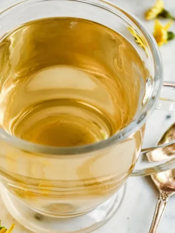 Clear glass mug filled with golden roasted dandelion root tea beside a spoon and scattered dandelion flowers.