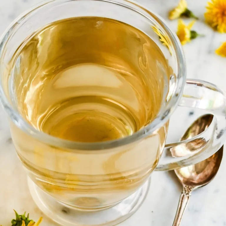 Clear glass mug filled with golden roasted dandelion root tea beside a spoon and scattered dandelion flowers.