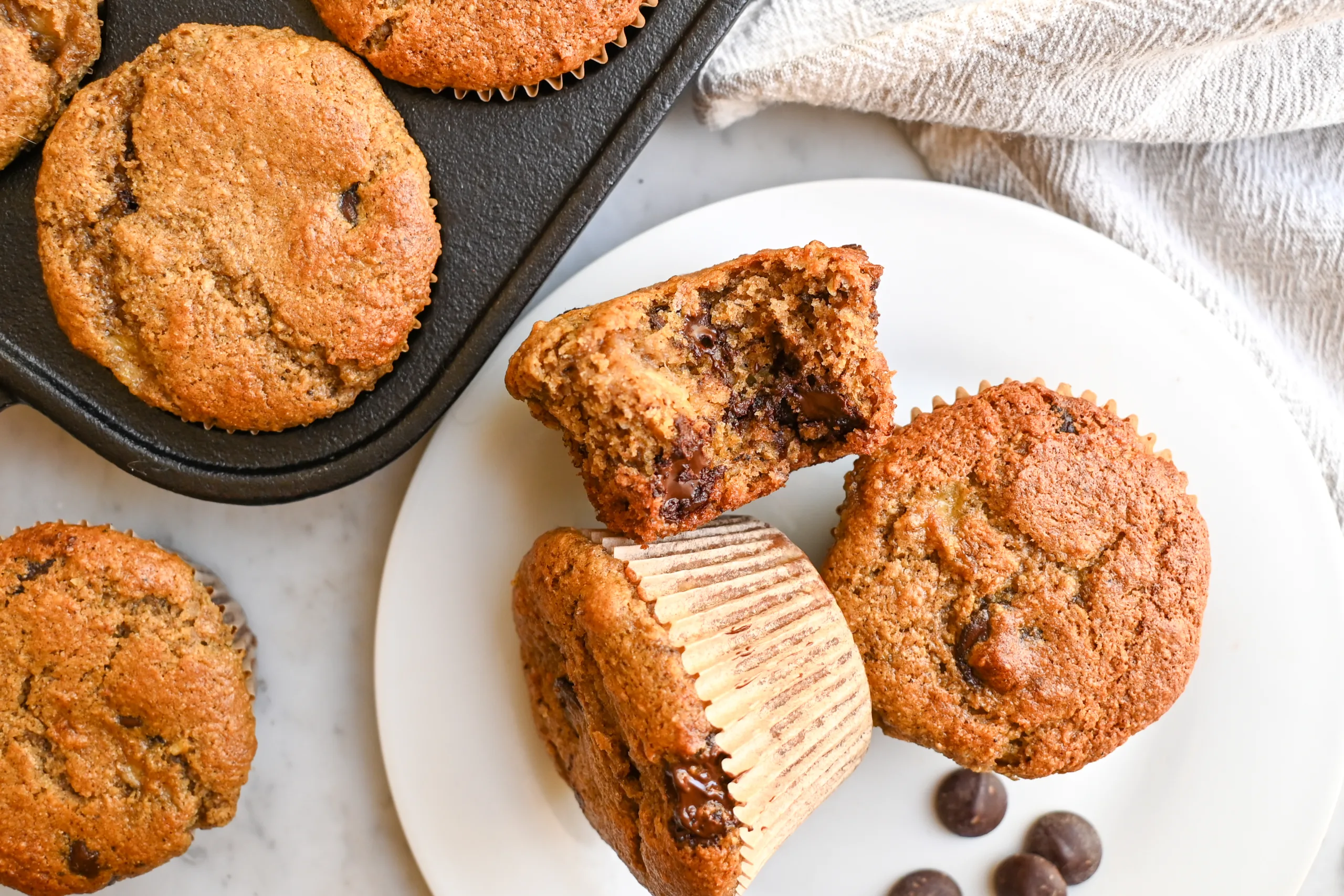 Homemade whole wheat banana muffins with chocolate chips on a plate beside a muffin pan.