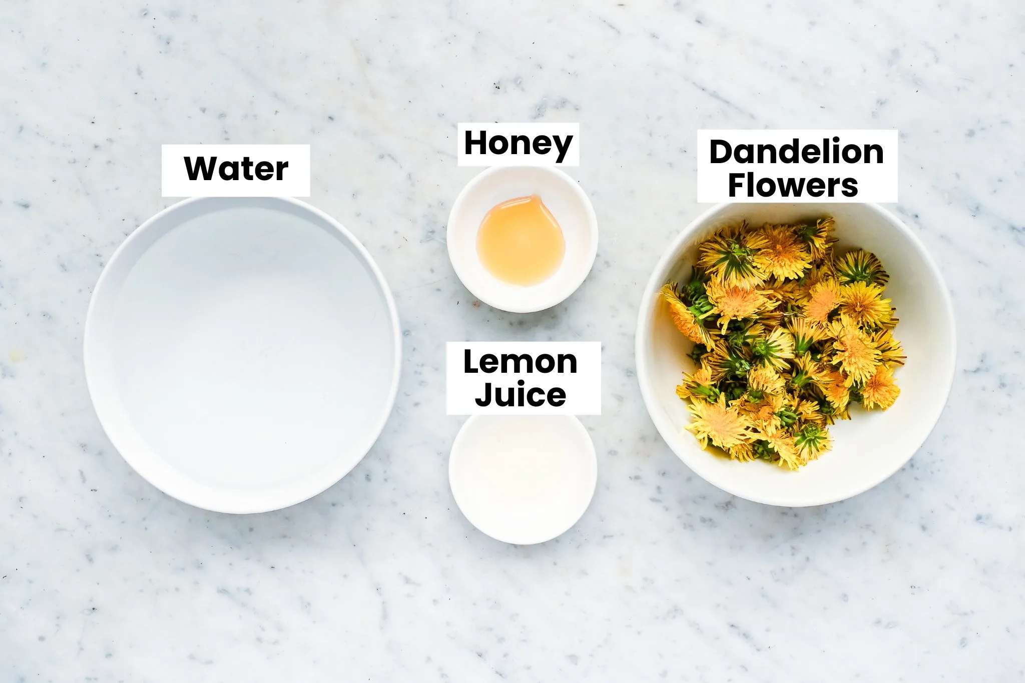 Ingredients for fresh dandelion tea with dandelion flowers, water, honey, and lemon juice on a marble counter.