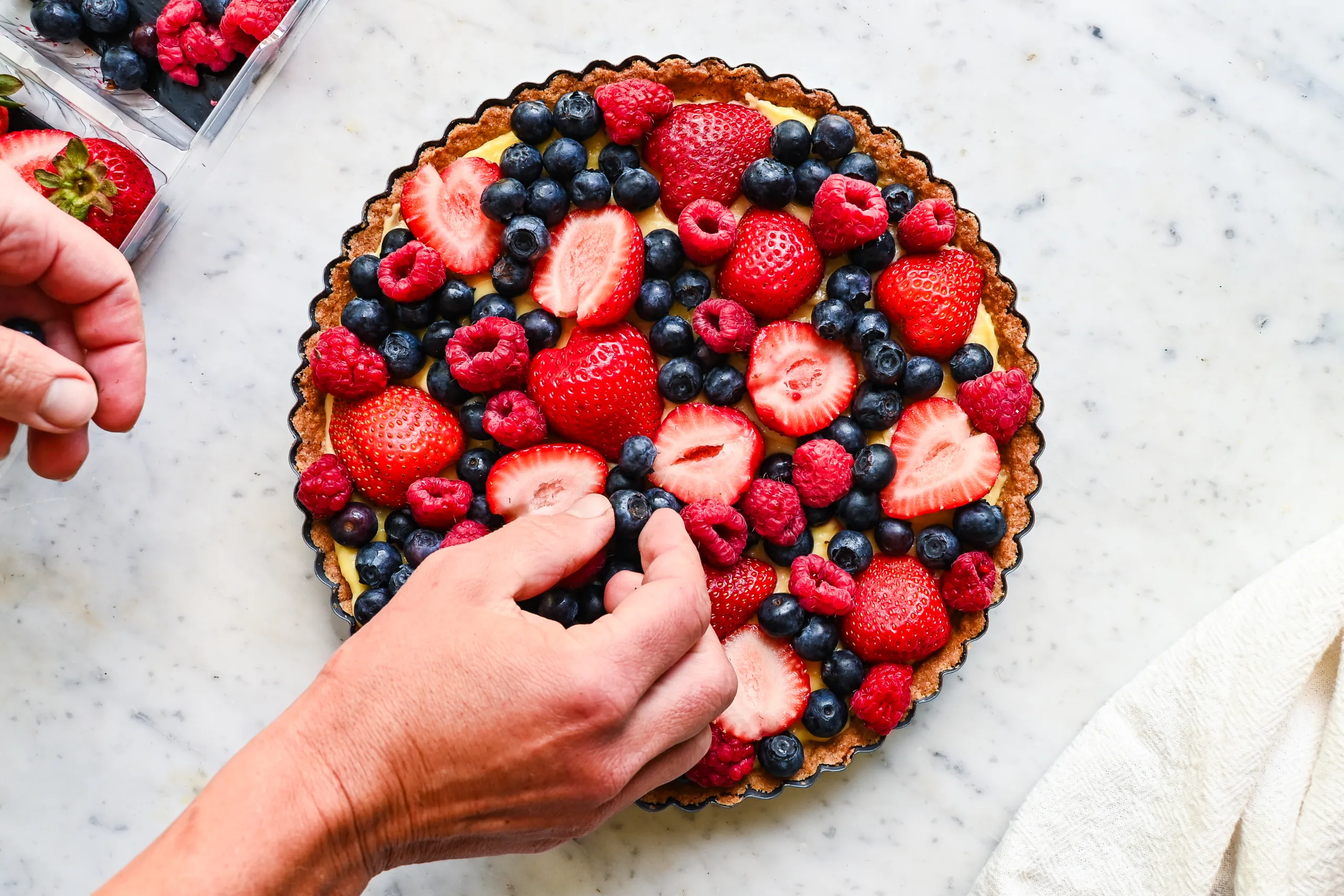 Fresh strawberries, raspberries, and blueberries being arranged on top of a vanilla cream tart.