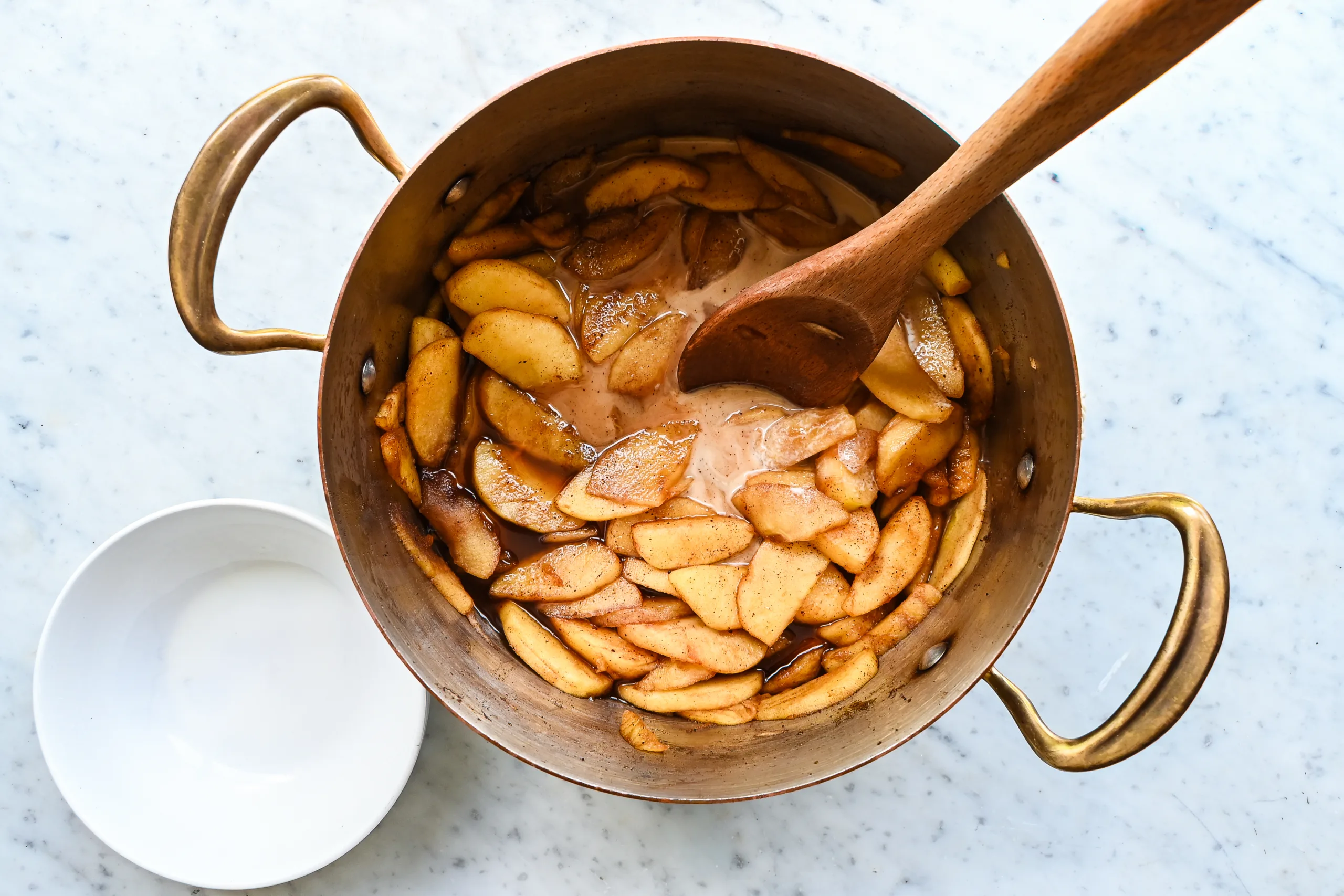 Cornstarch slurry being added to the apple slices that are cooking in cinnamon sugar syrup with a wooden spoon.