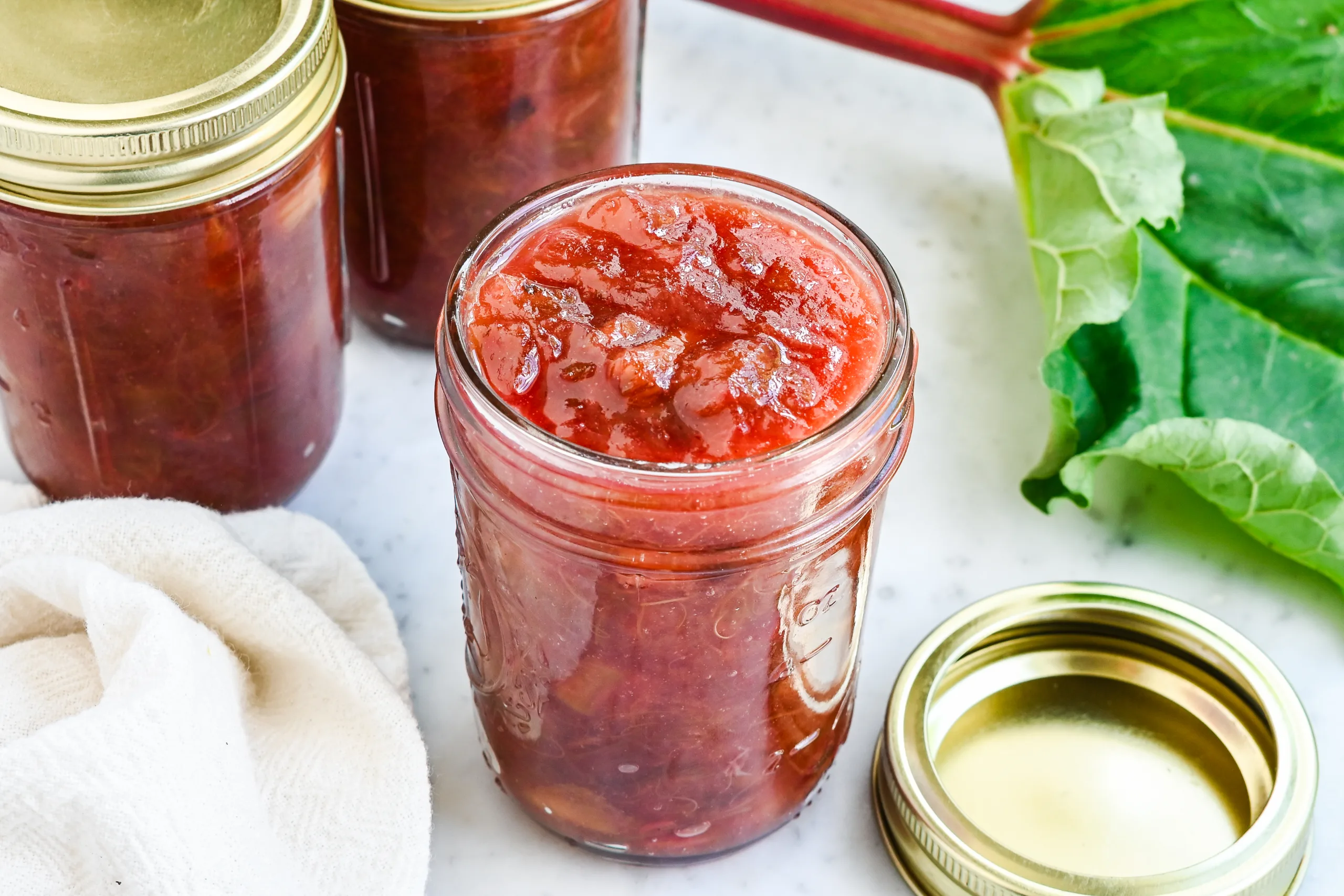 Open glass jar filled with homemade rhubarb jam beside rhubarb stalks and leaves.