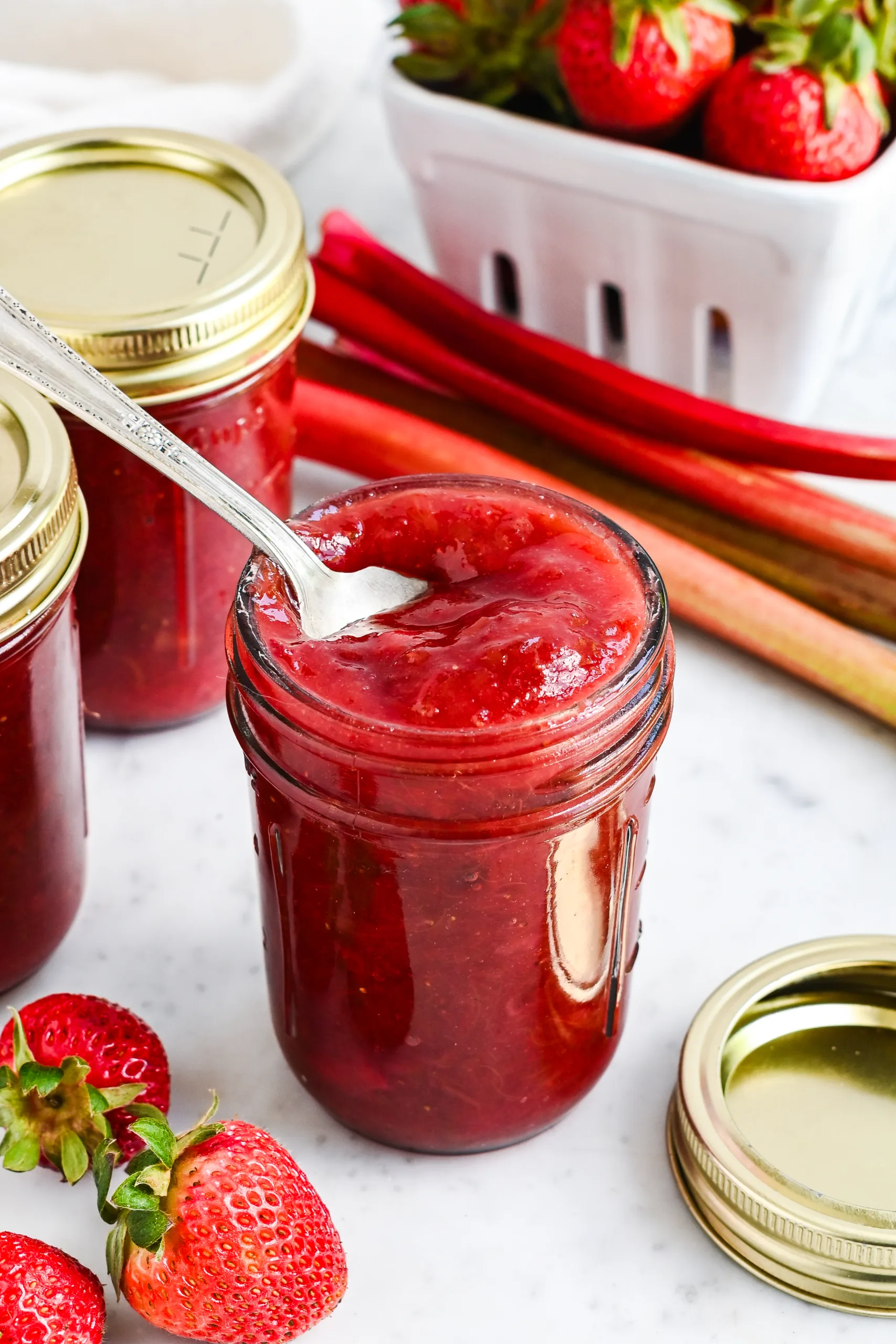Spoon dipped into a jar of homemade strawberry rhubarb jam with strawberries and rhubarb nearby.