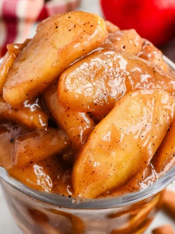 This close-up of homemade apple pie filling is the best choice for the feature image because it shows the finished recipe clearly. The glossy cinnamon-coated apples look rich and thick, and the fresh apples and cinnamon sticks in the background help readers instantly understand what the recipe is.