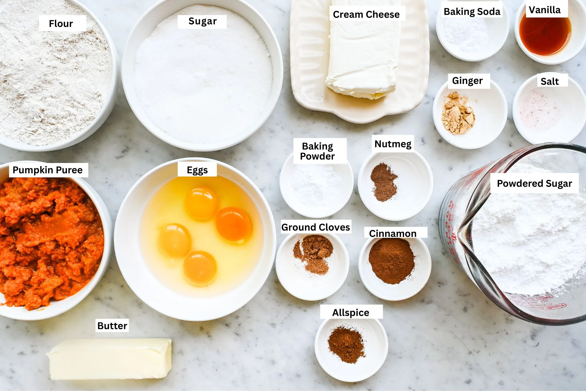 Pumpkin bars ingredients arranged on a marble counter with pumpkin purée, eggs, flour, sugar, butter, cream cheese, powdered sugar, vanilla, and spices.
