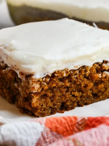 Pumpkin bar with cream cheese frosting served on a white plate with a plaid towel, fork, and frosted bars in the background.