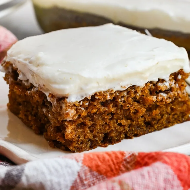 Pumpkin bar with cream cheese frosting served on a white plate with a plaid towel, fork, and frosted bars in the background.