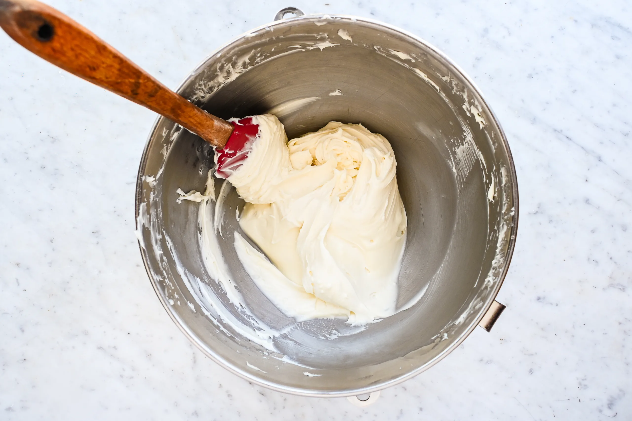 Cream cheese frosting mixed in a stand mixer bowl with a red spatula.