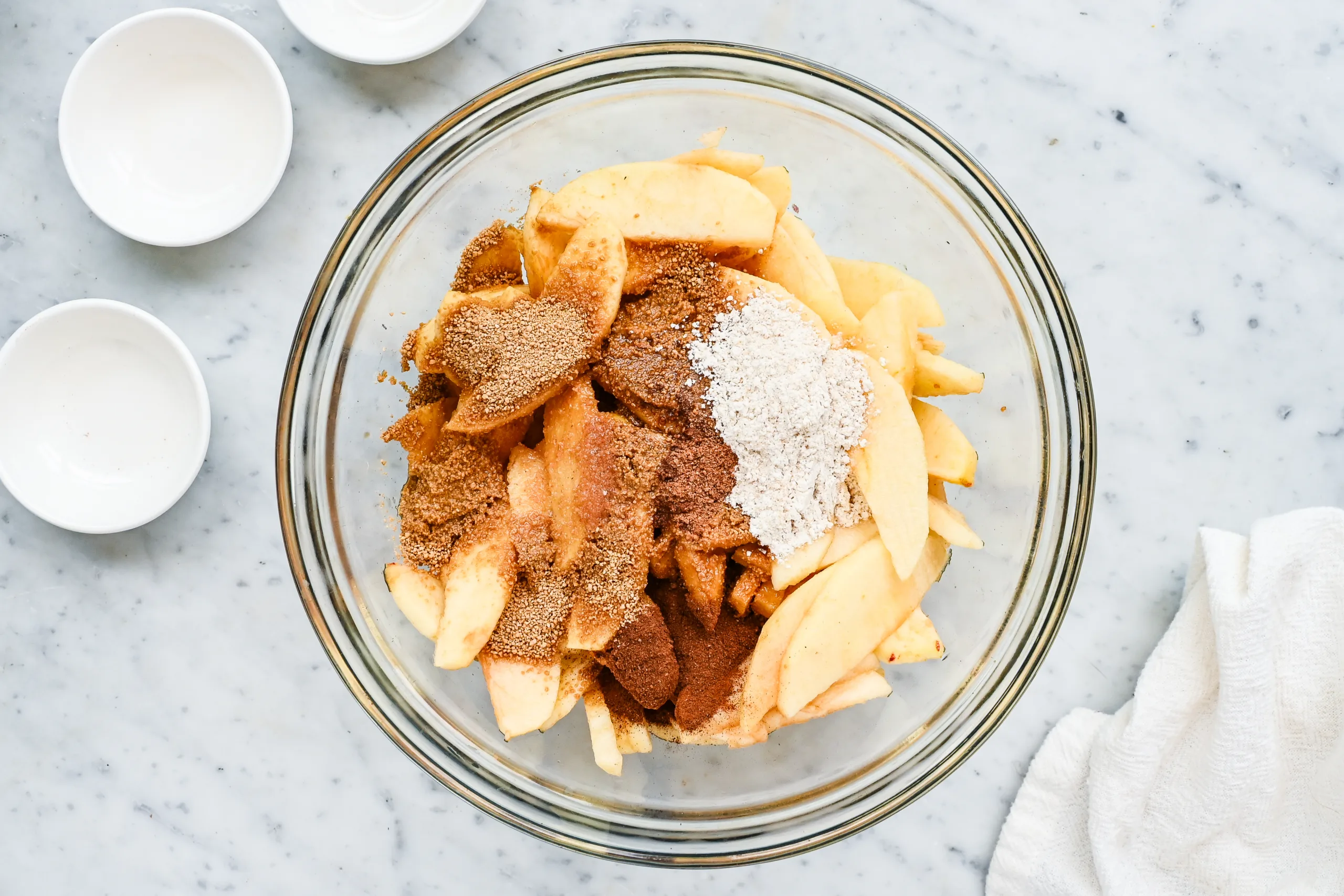 Sliced apples with brown sugar, flour, cinnamon, nutmeg, salt, lemon juice, and vanilla in a glass mixing bowl.
