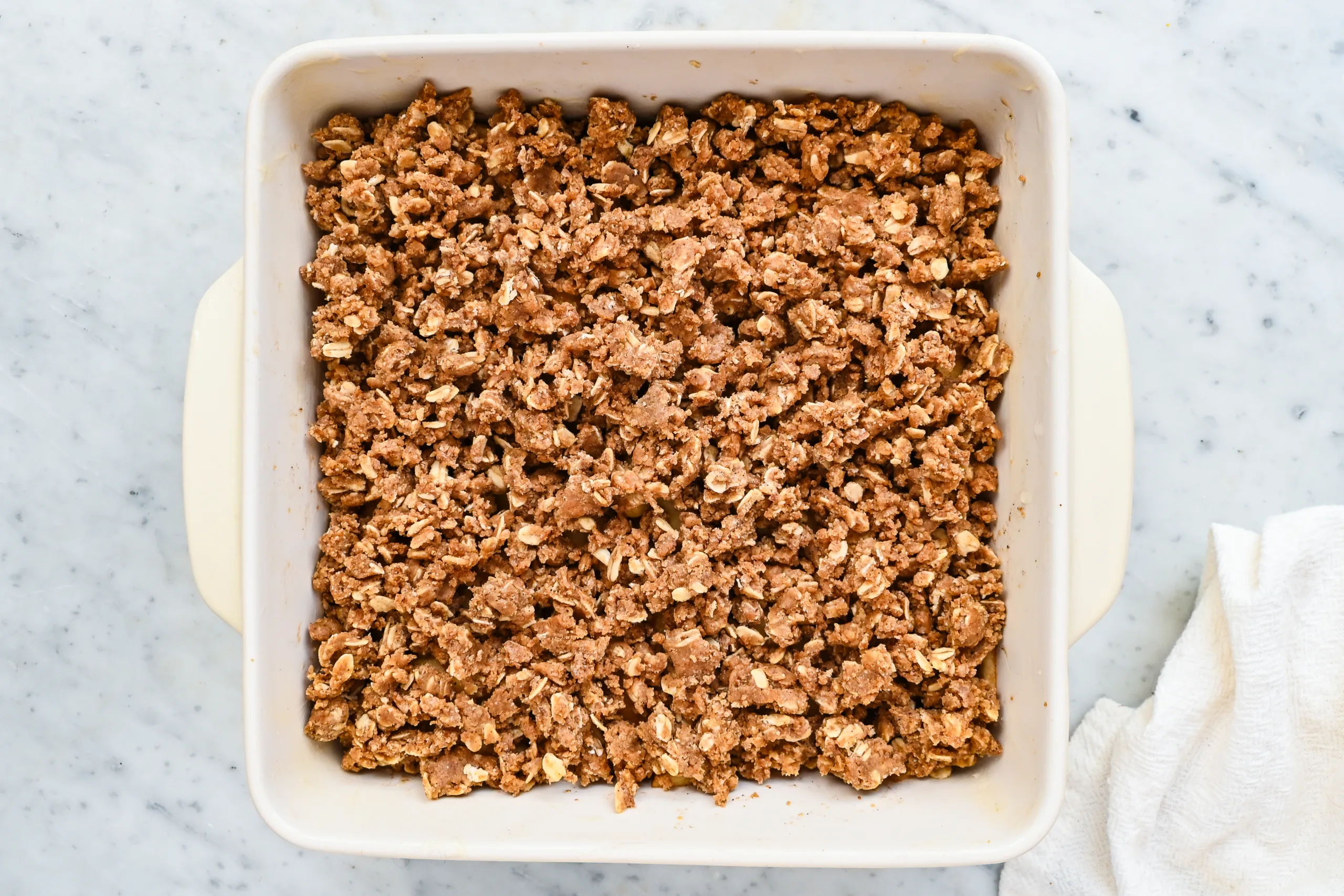 Sourdough oat crumble topping spread over cinnamon apple filling in a white baking dish before baking.