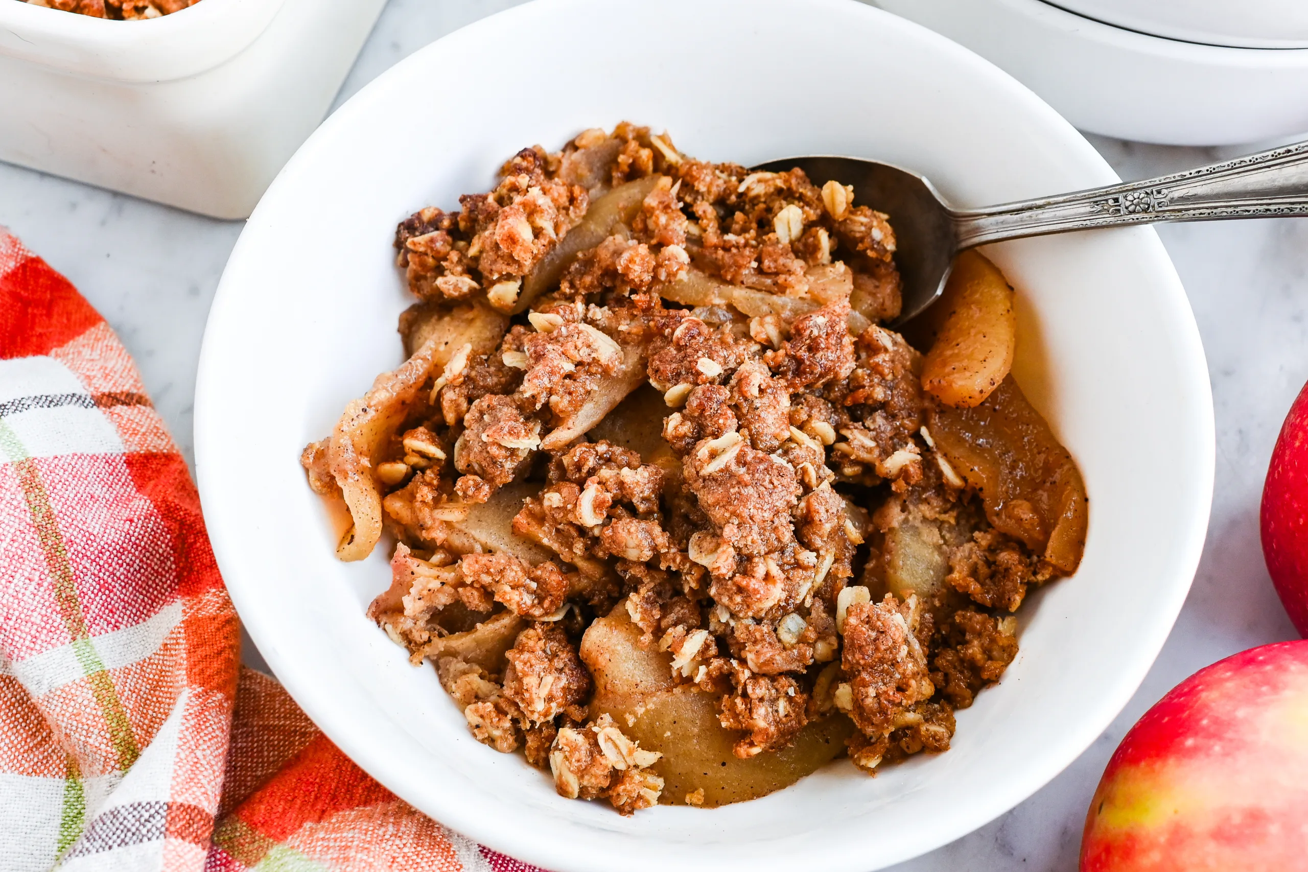 Sourdough apple crisp with oat crumble topping served in a white bowl with a spoon, plaid towel, and fresh apples nearby.