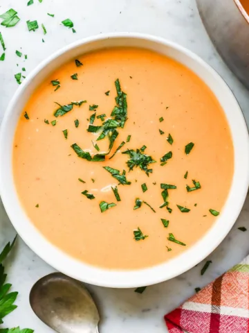 Creamy butternut pumpkin sweet potato soup in a white bowl with parsley, a copper pot, plates, and a plaid towel nearby.