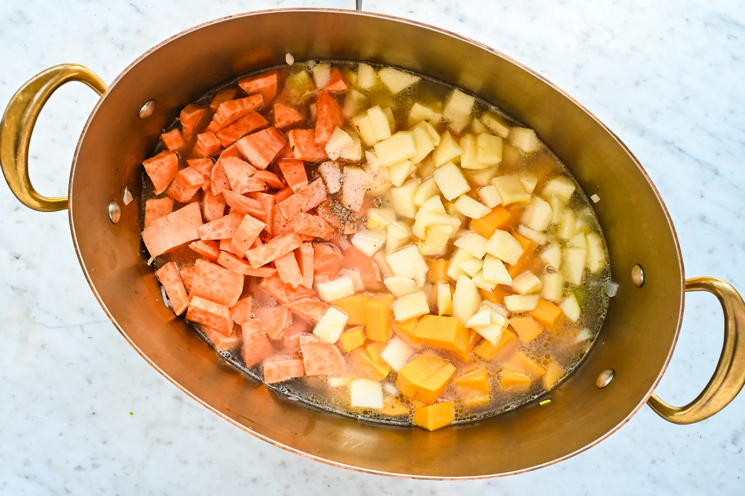 Butternut pumpkin, sweet potatoes, apple, and broth added to a copper pot for soup.