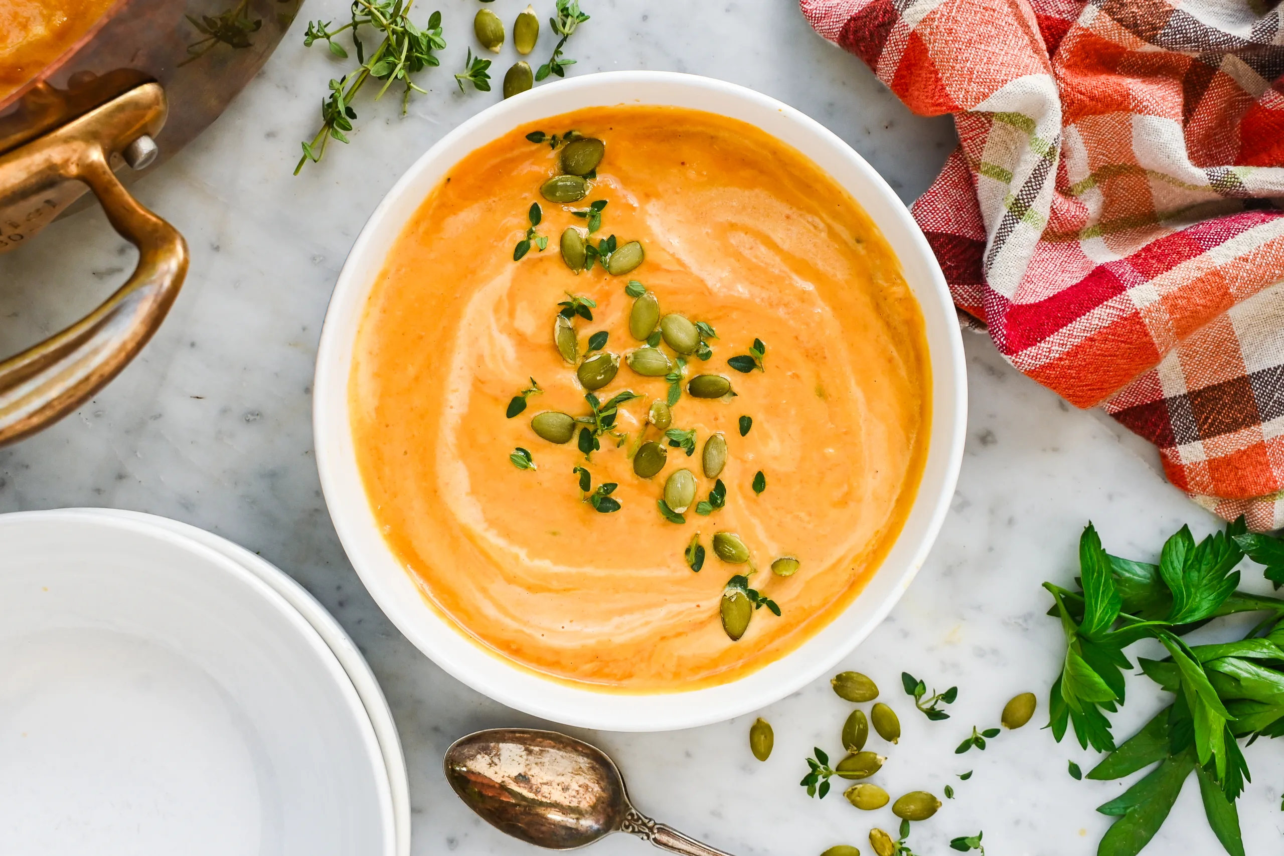A bowl of creamy pumpkin soup with pumpkin seeds, fresh thyme, and cream swirls, served beside a copper pot and fall plaid towel.