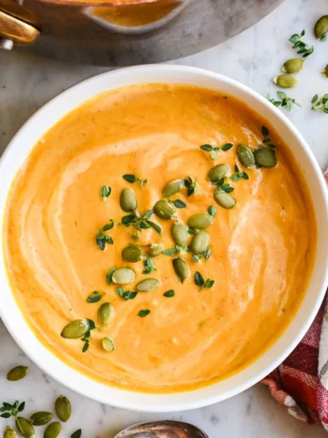 Creamy pumpkin soup in a white bowl topped with pumpkin seeds and fresh thyme, with a copper pot and plaid towel nearby.