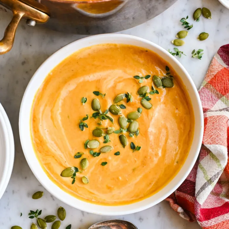 Creamy pumpkin soup in a white bowl topped with pumpkin seeds and fresh thyme, with a copper pot and plaid towel nearby.