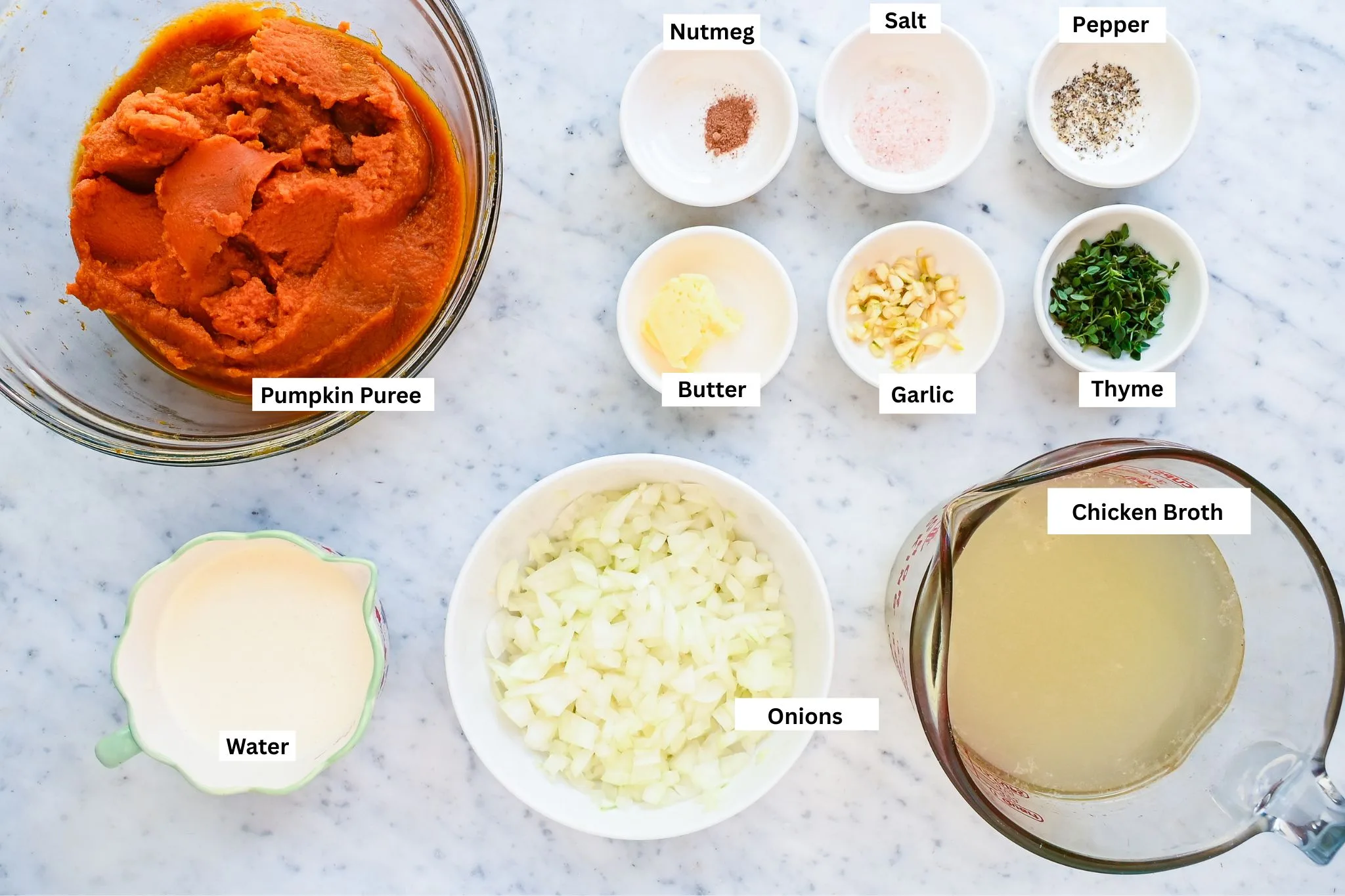 Pumpkin soup ingredients arranged on a marble counter, including pumpkin purée, chopped onion, broth, cream, garlic, butter, thyme, salt, pepper, and nutmeg.