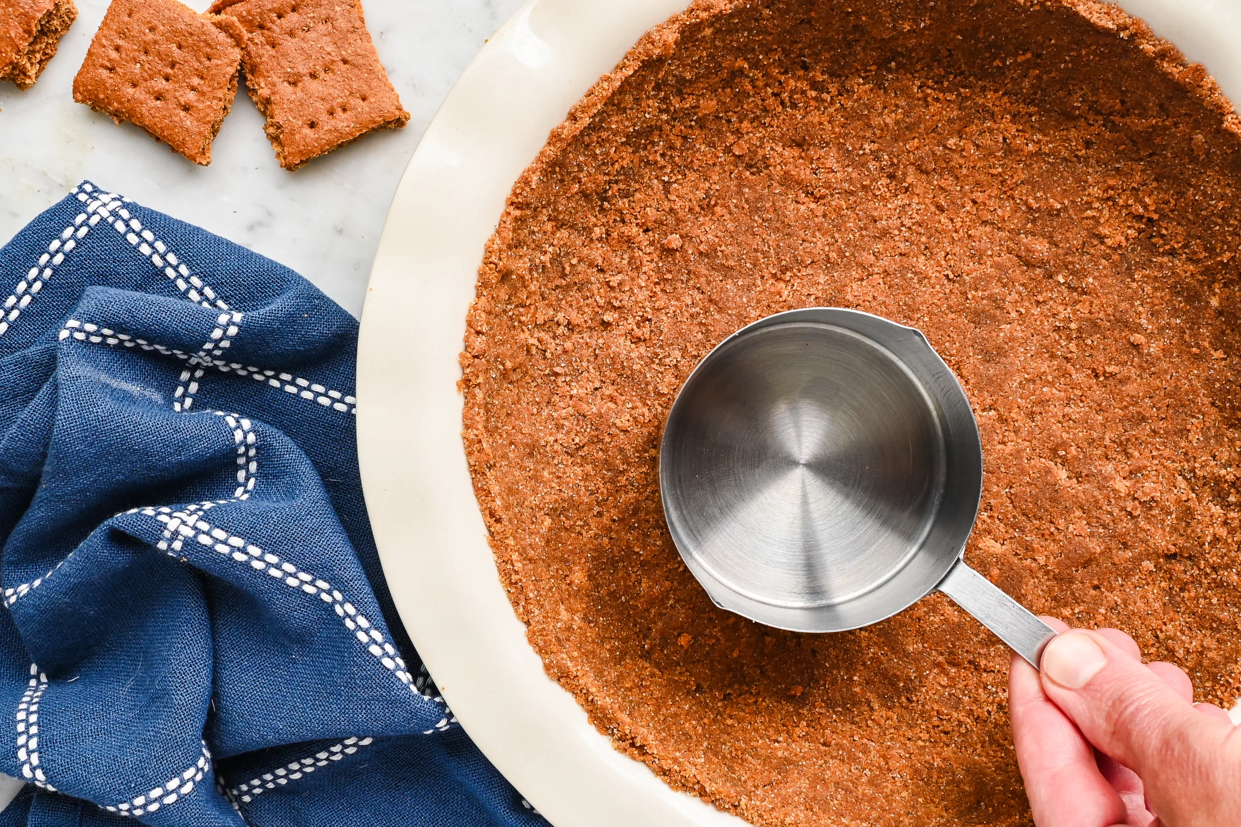 Close-up of a graham cracker pie crust being shaped with a metal measuring cup.