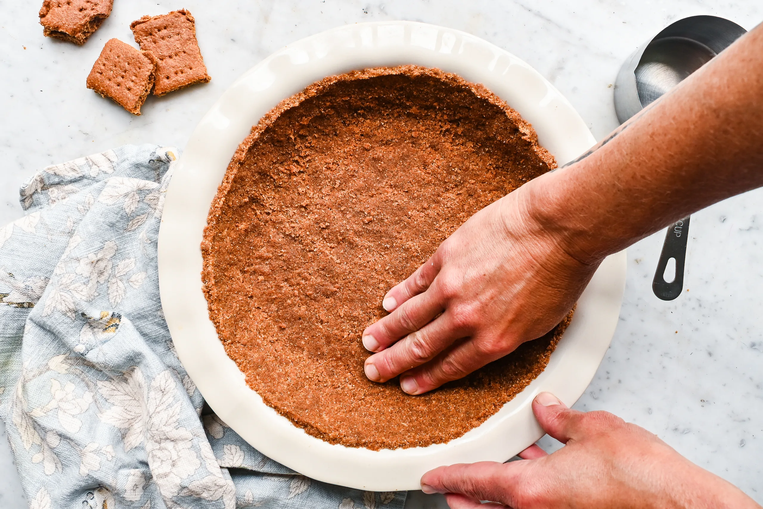 Hands pressing graham cracker crumbs into a white pie plate to form a crust.