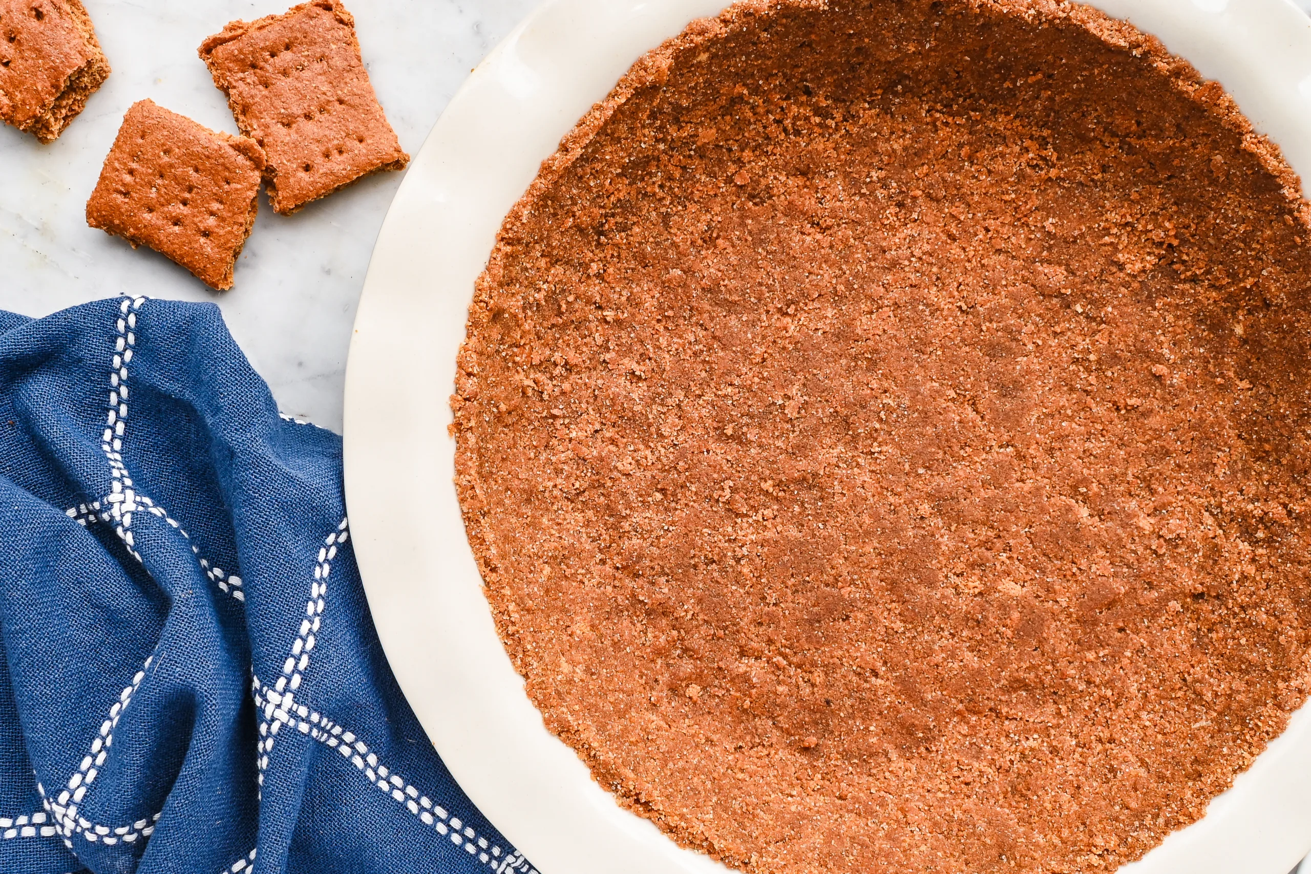 Graham cracker pie crust pressed into a white pie plate with graham crackers and a blue towel nearby.