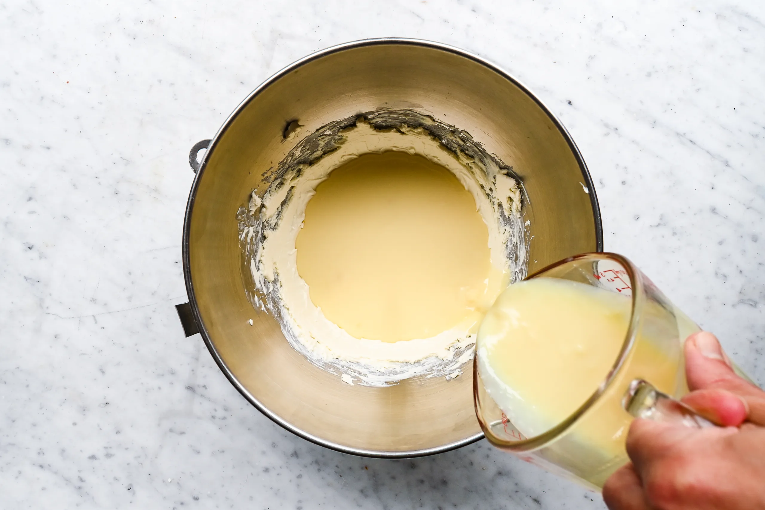 Sweetened condensed milk being poured into cream cheese in a stand mixer bowl.