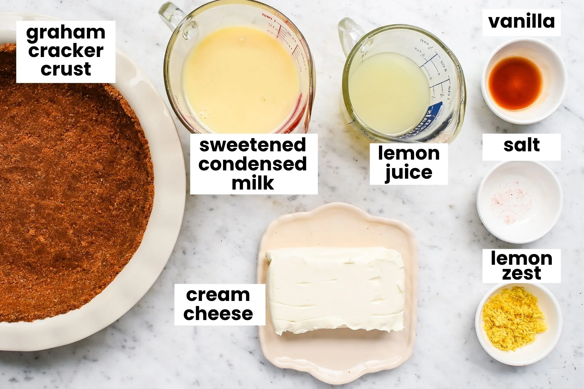 No-bake lemon icebox pie ingredients arranged on a marble counter, including graham cracker crust, cream cheese, sweetened condensed milk, lemon juice, lemon zest, vanilla, and salt.