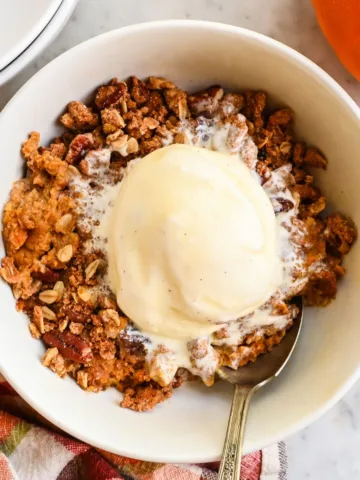 Pumpkin crisp served warm in a white bowl with vanilla ice cream, oat pecan topping, cinnamon sticks, a pumpkin, and a plaid towel nearby.